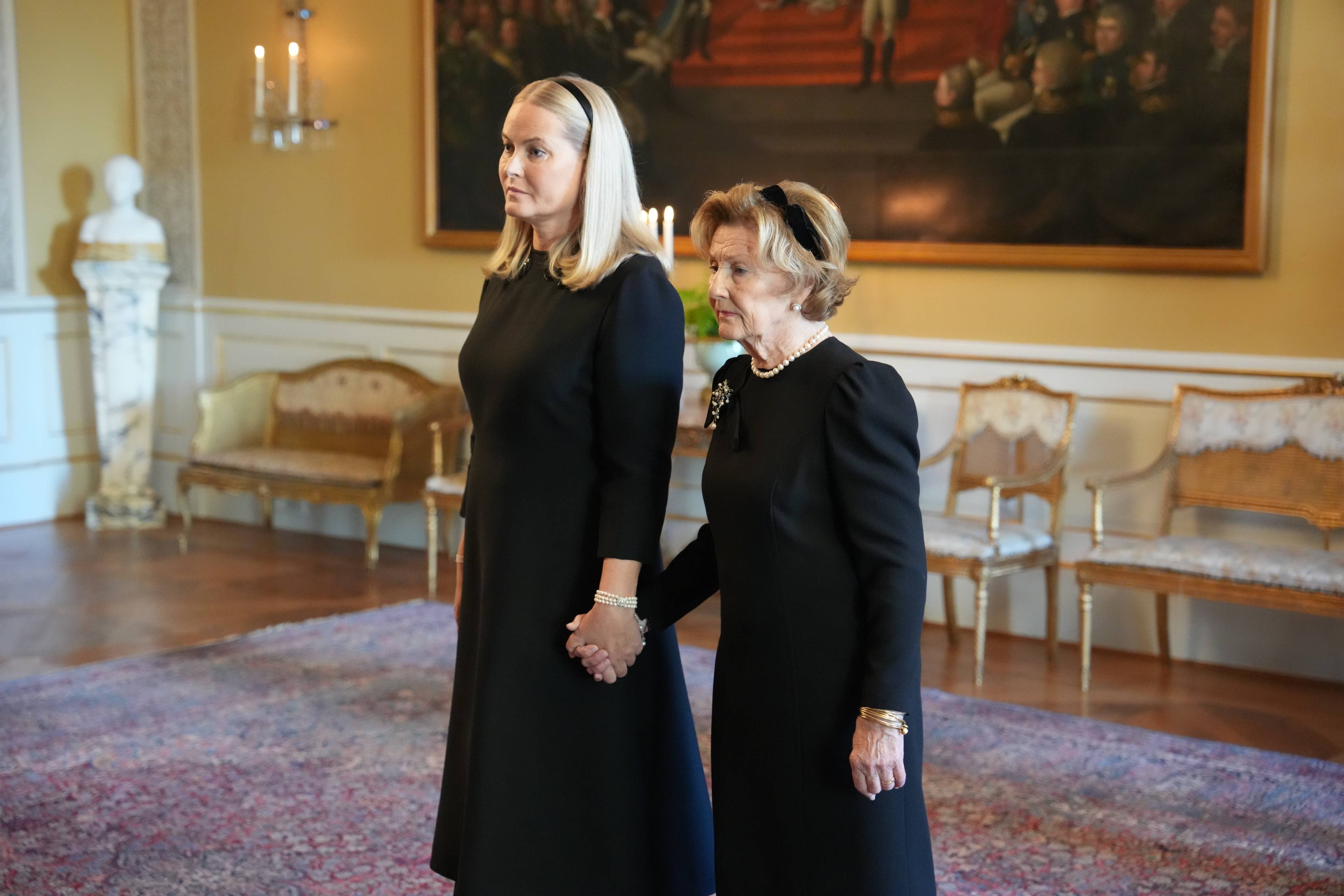 Dronning Mette-Marit og dronning Sonja holder hverandre i hendene i en stue på Slottet. (Bildebeskrivelsen er laget av en KI-tjeneste)