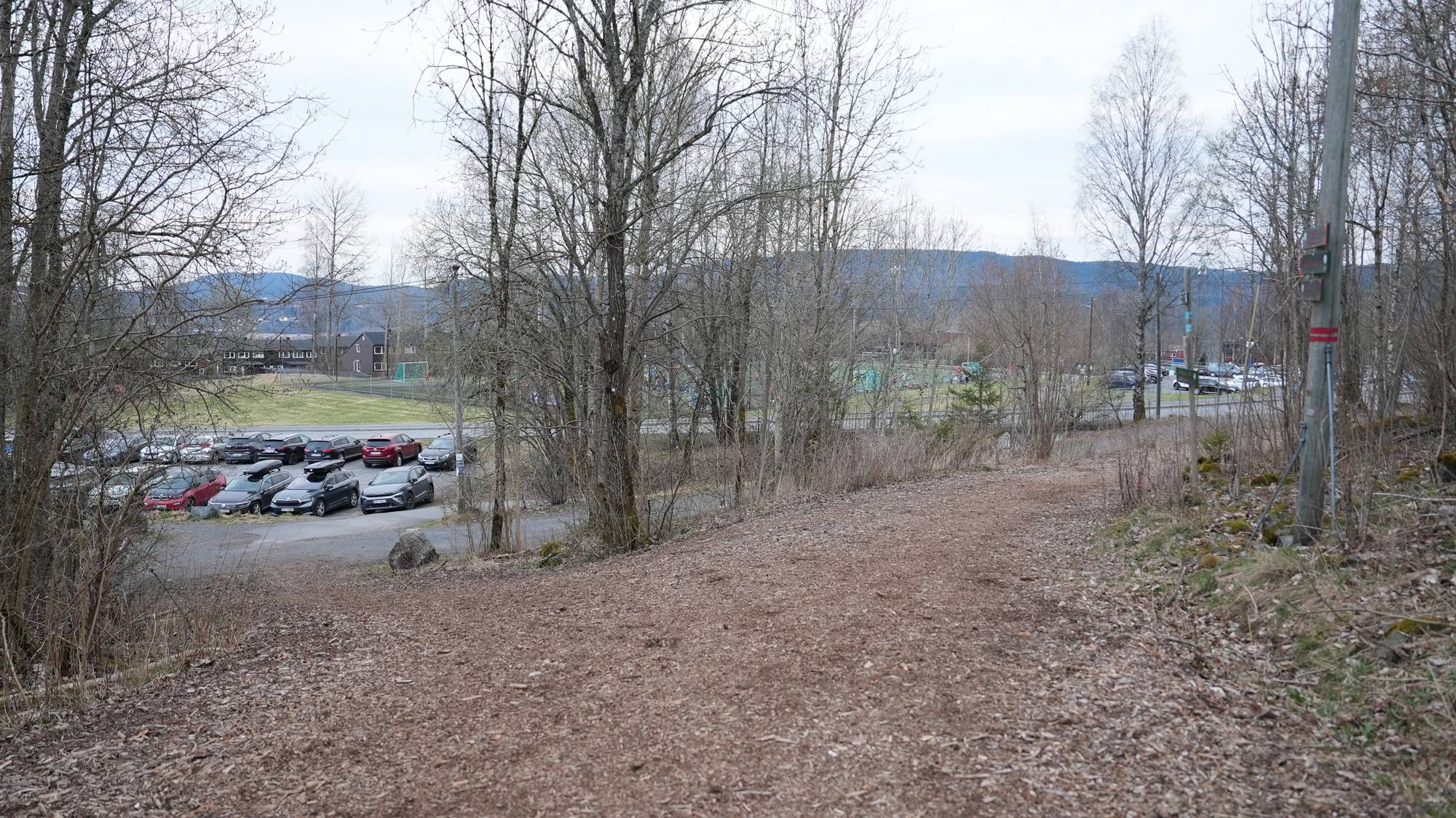Parkerte biler står på en grusbane. I bakgrunnen ses bygninger og en idrettsplass. Bildet er tatt fra en sti i skogen. Det er en grå og skyete dag. (Bildebeskrivelsen er laget av en KI-tjeneste)