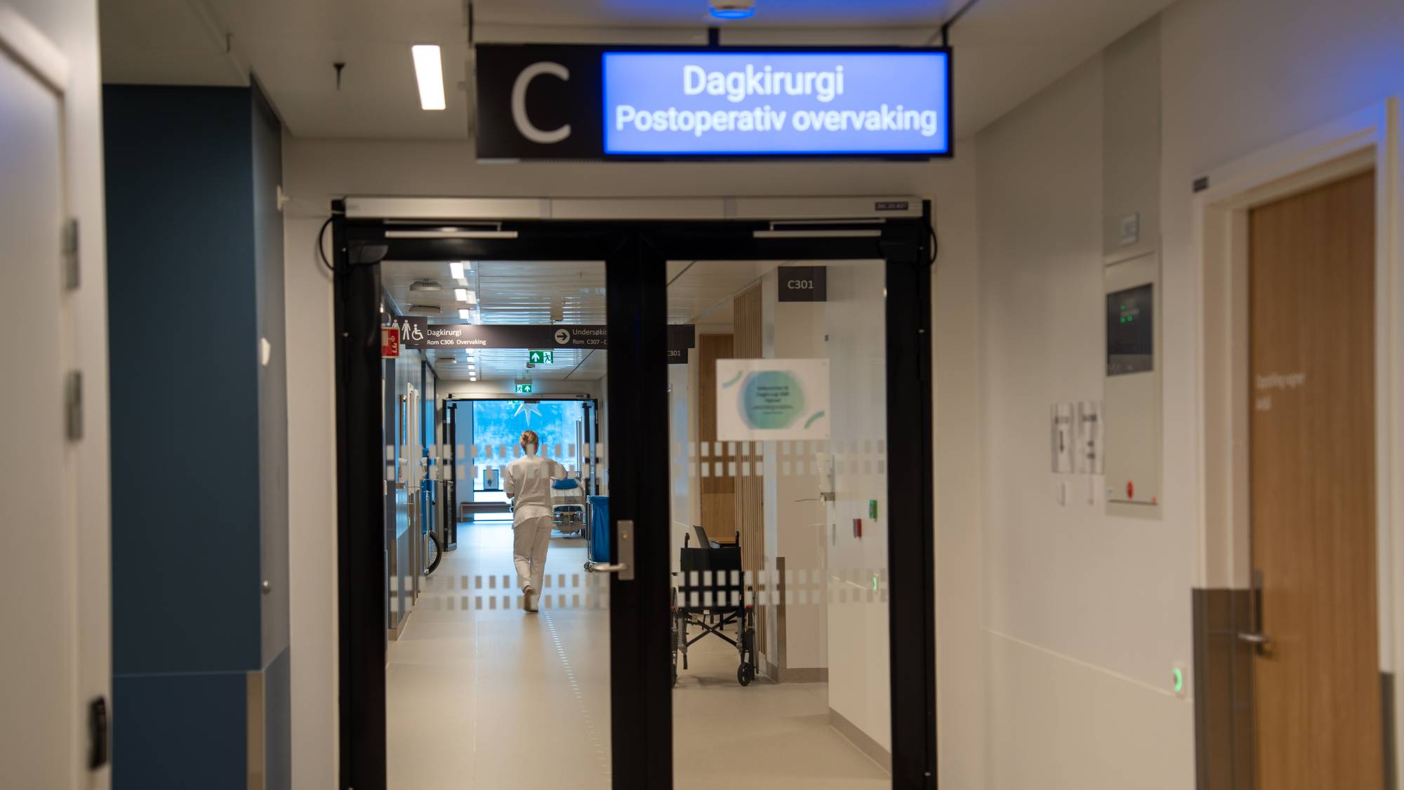Sykehuskorridor ved dagkirurgi med skilt og en helsearbeider i hvit uniform som går gjennom døren. (Bildebeskrivelsen er laget av en KI-tjeneste)