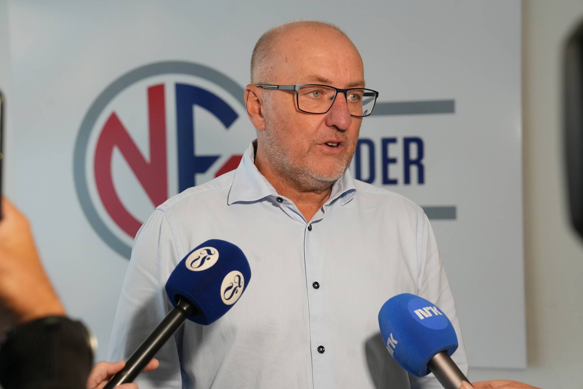 En mann med briller snakker til mikrofoner foran en logo fra Norsk Fysioterapeutforbund. (Bildebeskrivelsen er laget av en KI-tjeneste)