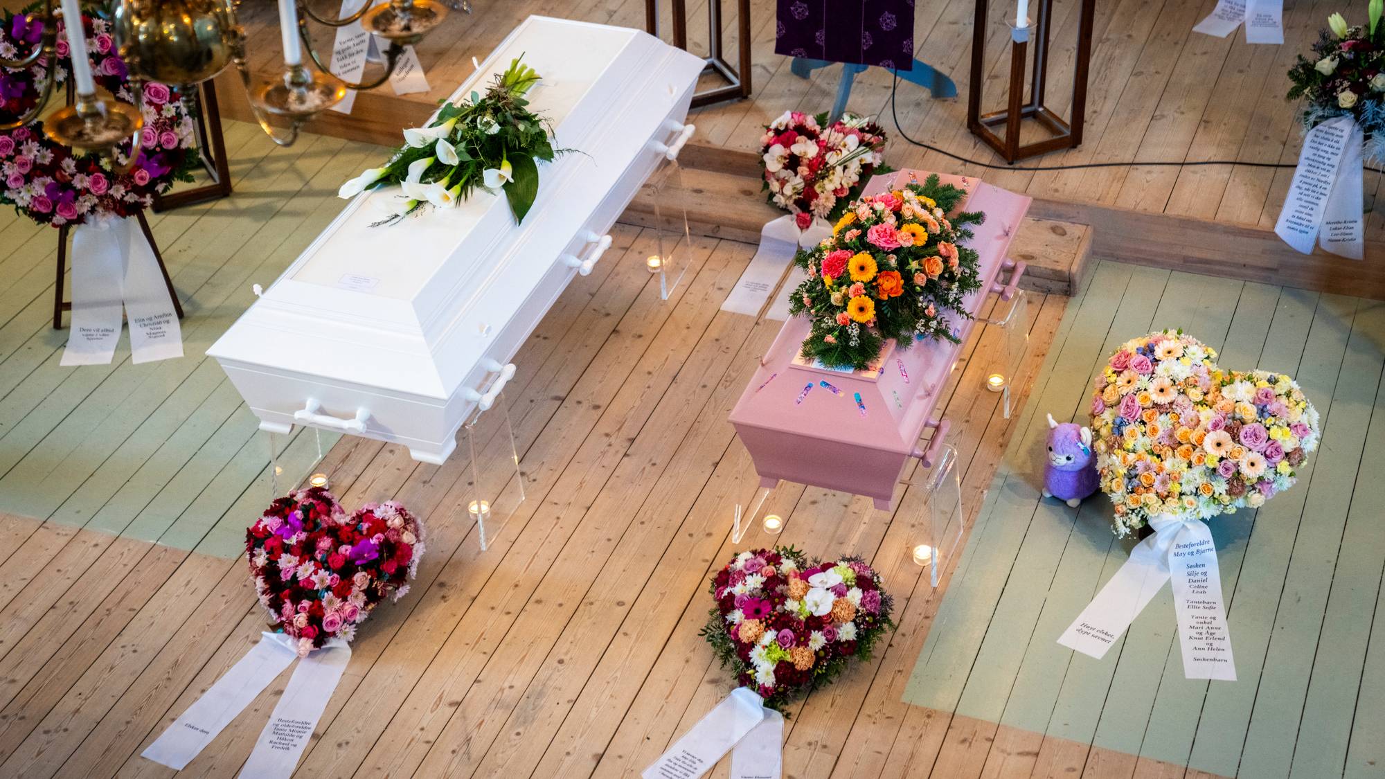 To kister, en hvit og en rosa, er plassert side om side i et rom. Rundt kistene ligger det flere blomsterdekorasjoner, blant annet i hjerteform. (Bildebeskrivelsen er laget av en KI-tjeneste)