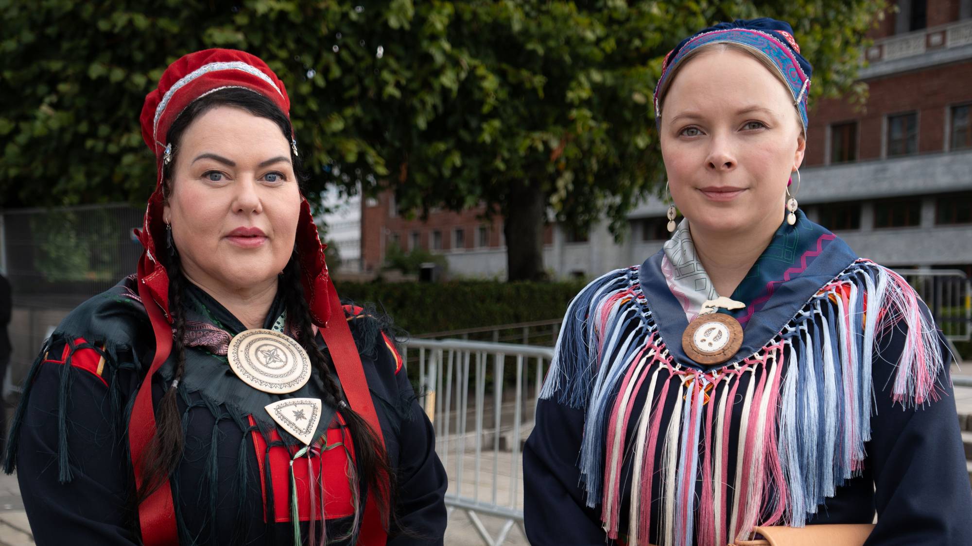 To kvinner i tradisjonelle samiske kofter står utendørs foran en murbygning. (Bildebeskrivelsen er laget av en KI-tjeneste)