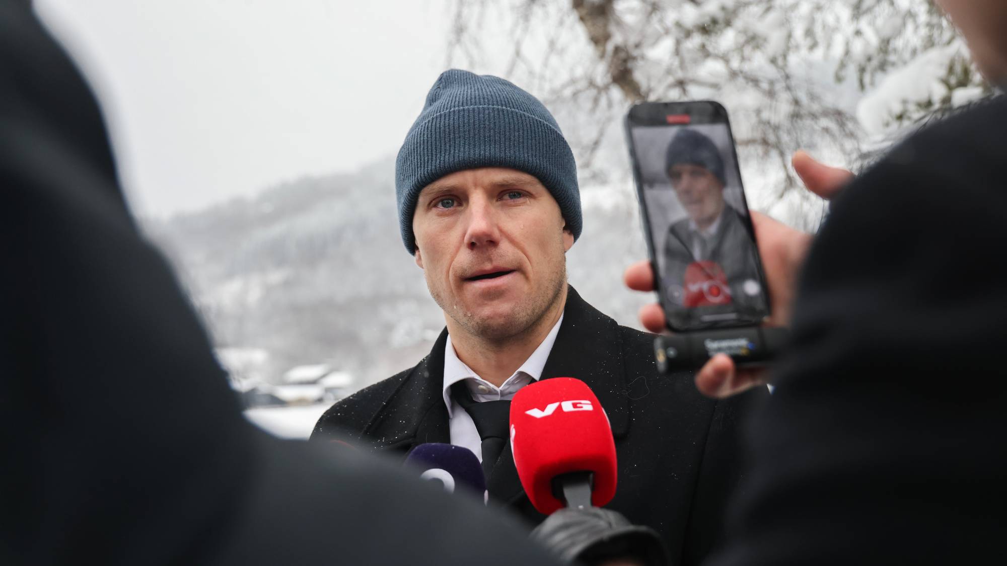 En mann i dress og lue står ute og blir intervjuet. Han holder en mikrofon, mens flere personer rundt ham tar bilder med mobiltelefoner. Omgivelsene er snødekte med trær og bakgrunn av fjell. Det ser ut til å være vinter, og lysforholdene er dempet. (Bildebeskrivelsen er laget av en KI-tjeneste)