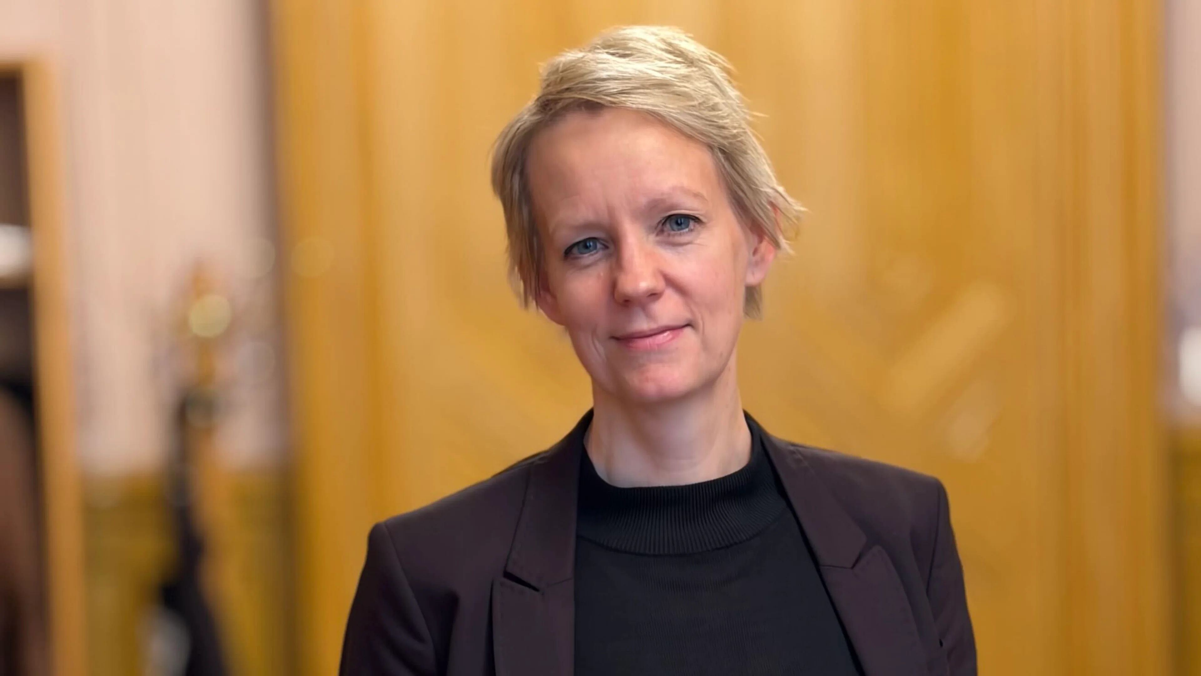 Ingrid fiskaa (SV) står på kontoret sitt på Stortinget, ikledd svart dress. Hun har kortklipt blondt hår.
