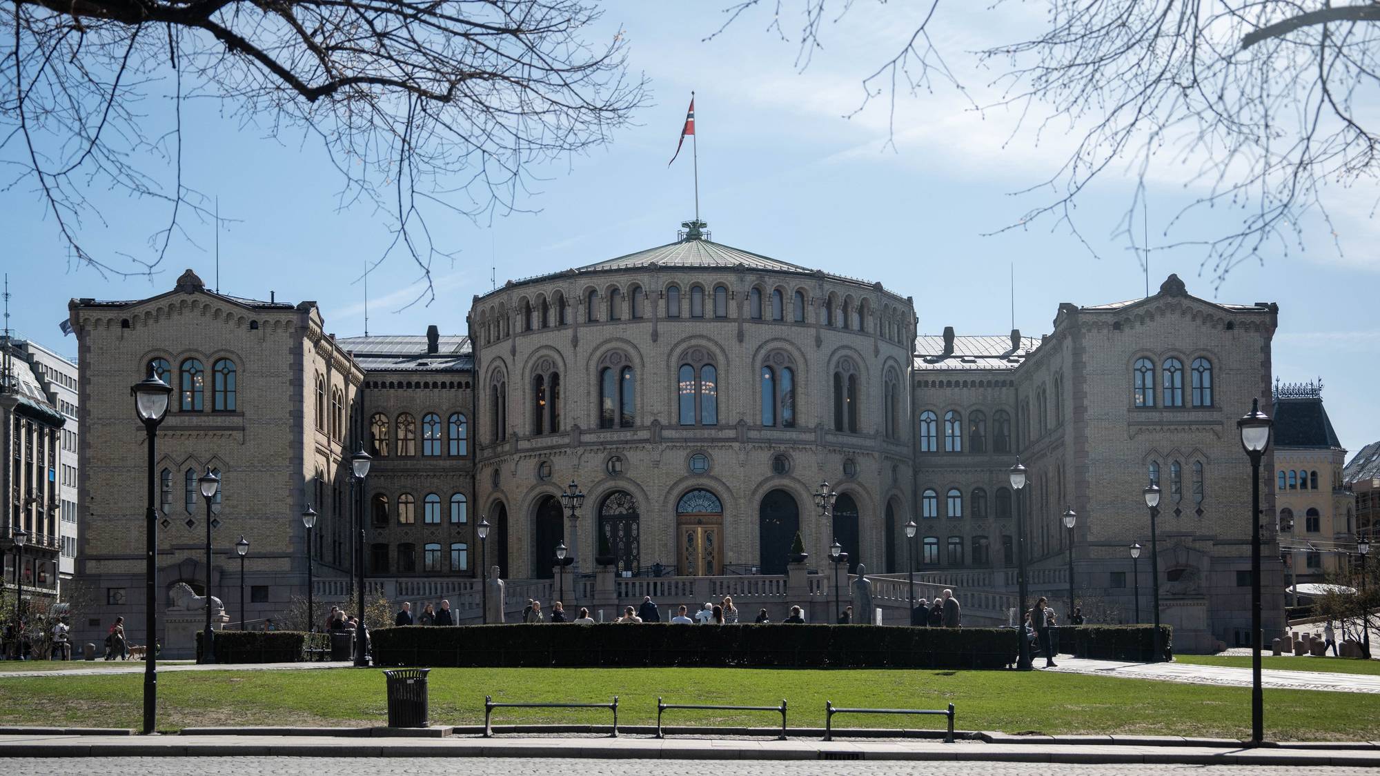 Det norske Stortinget er avbildet i sollys. Bygningen har gul murstein og et rundt midtparti. Utenfor sitter en gruppe mennesker på en benk foran en plen. Forgrunnen viser et brosteinsbelagt fortau. (Bildebeskrivelsen er laget av en KI-tjeneste)