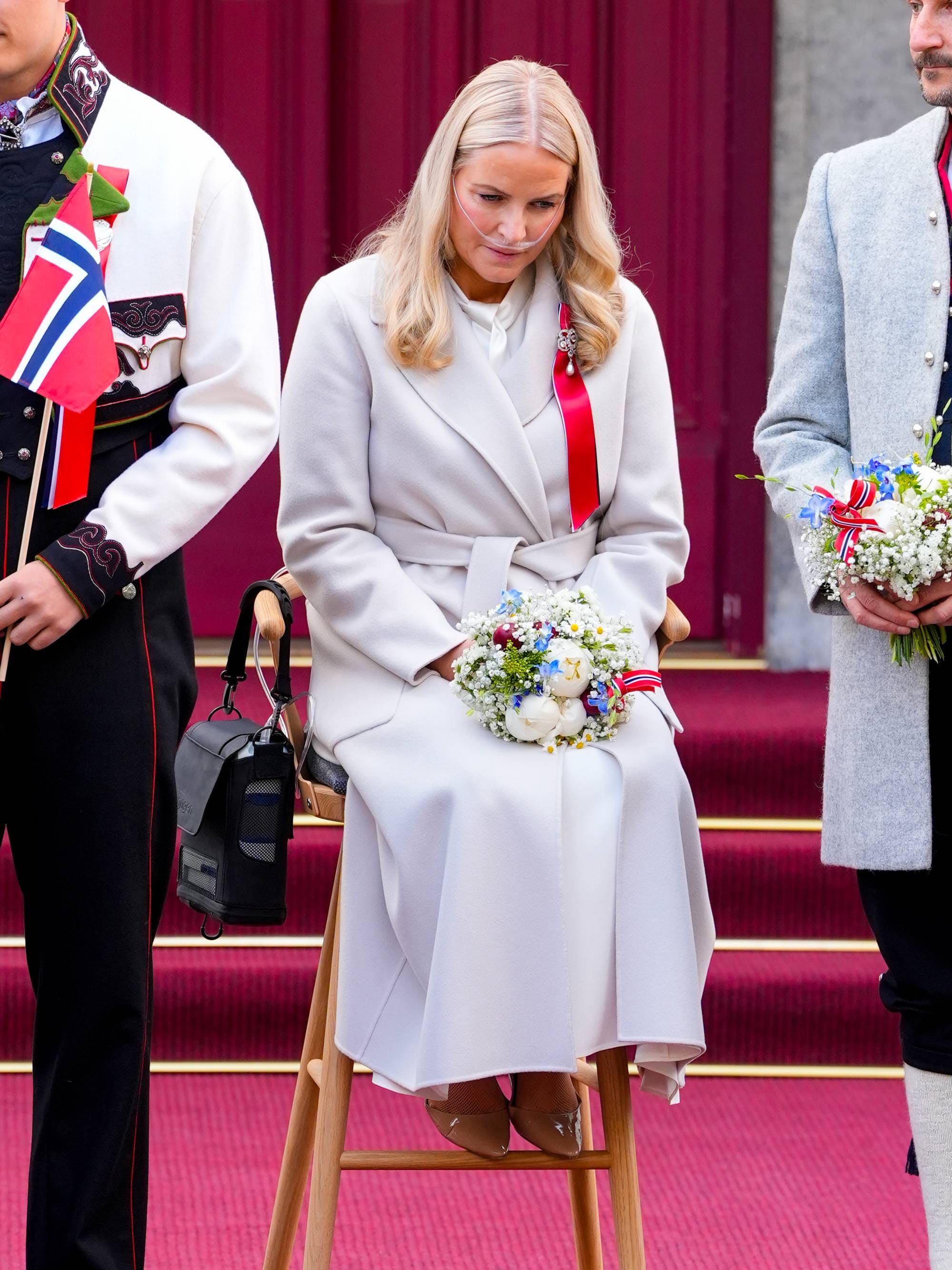 Kronprinsesse Mette-Marit sitter på en stol på 17. mai på Skaugum, hun ser ned og bruker et nesekateter