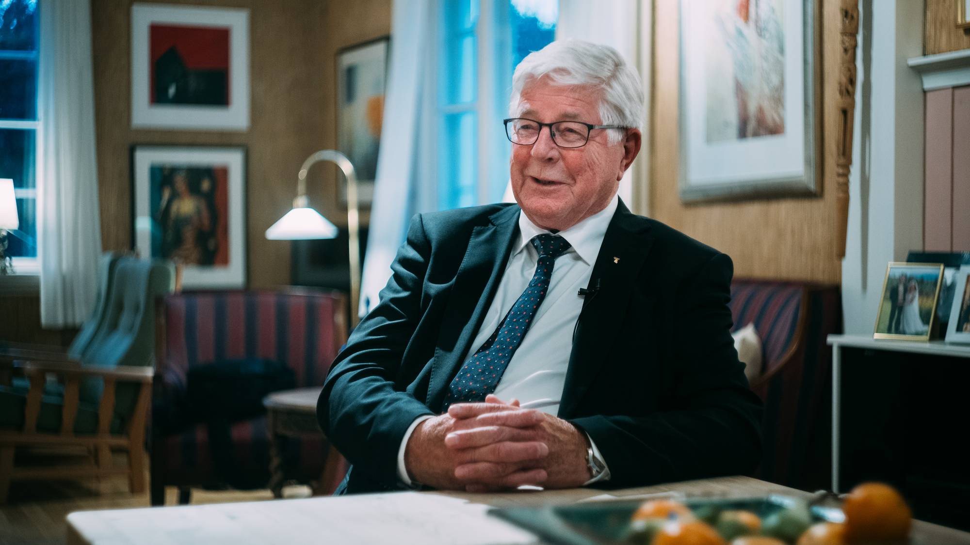 Eldre mann med briller og dress sitter ved et bord i en stue med bilder på veggene. (Bildebeskrivelsen er laget av en KI-tjeneste)