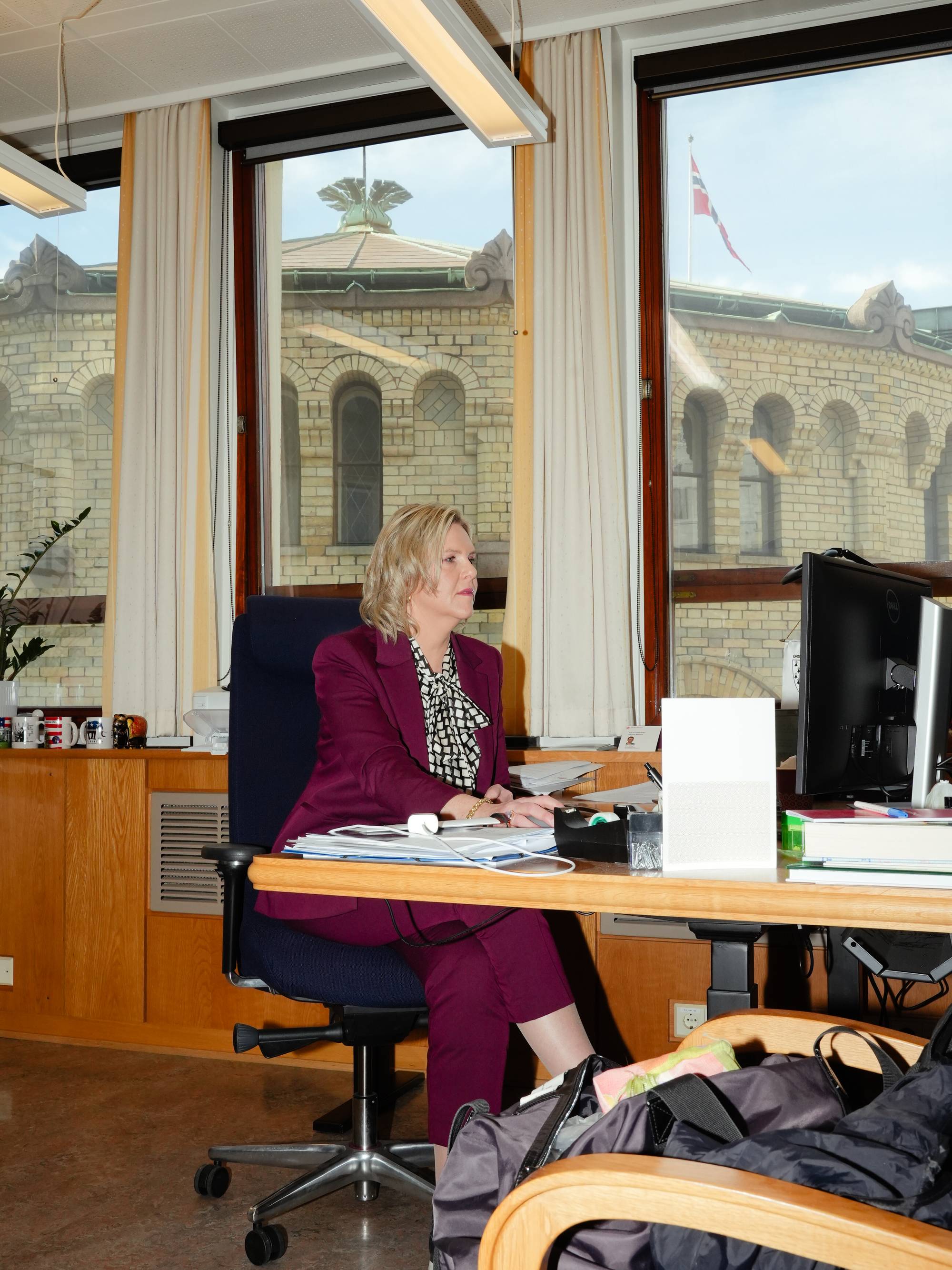 En kvinne i lilla dress sitter ved et skrivebord og ser på en dataskjerm. Bak henne er et vindu med utsikt mot en bygning med flagg. Hun har blondt hår og har papirer på skrivebordet. (Bildebeskrivelsen er laget av en KI-tjeneste)