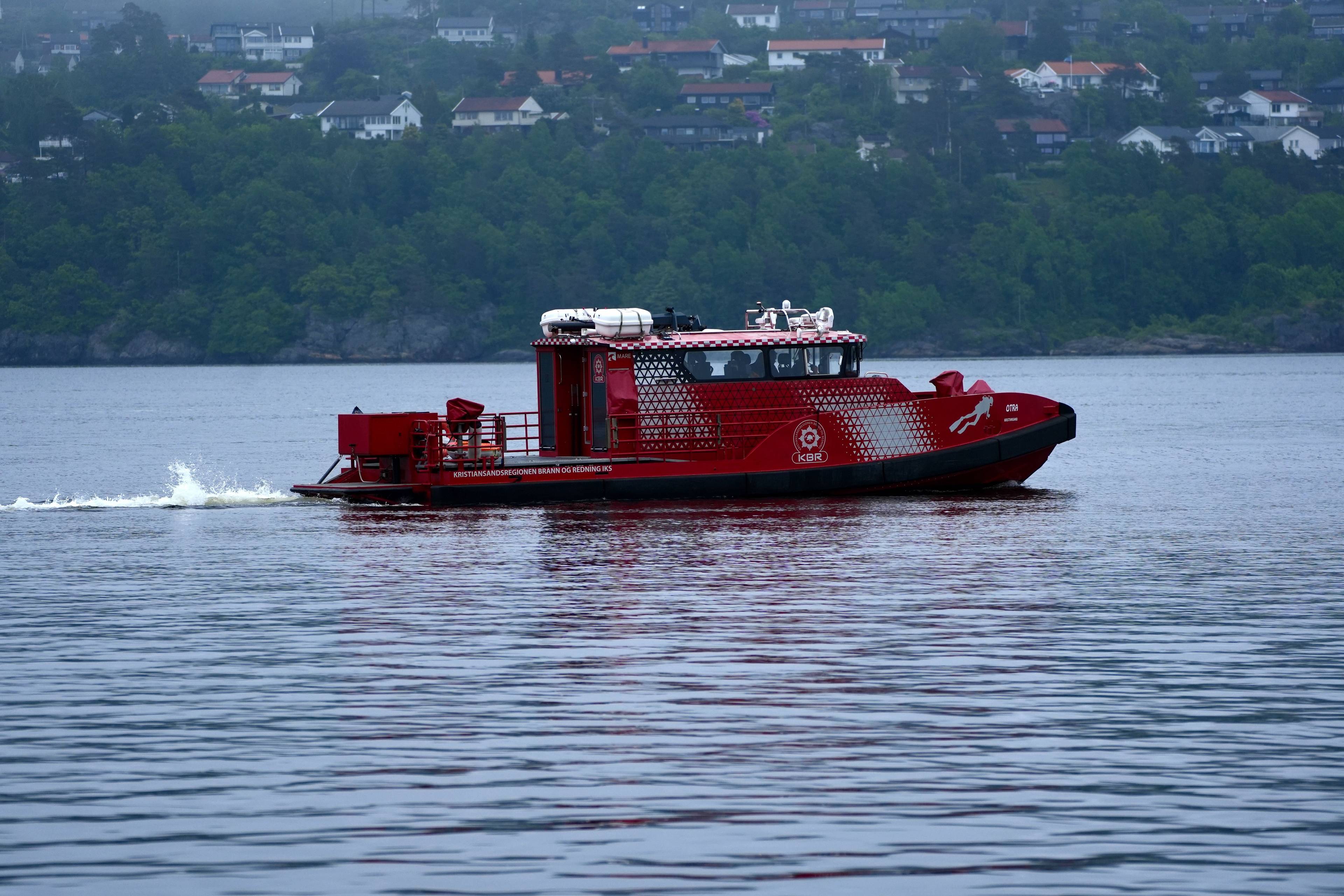Et rødt brannfartøy med logoen KBR seiler på vannet. Fartøyet har en lukket styrehus og et åpent dekk bak. Bak fartøyet er det en grønn ås med bebyggelse. Bildet er tatt fra siden. (Bildebeskrivelsen er laget av en KI-tjeneste)