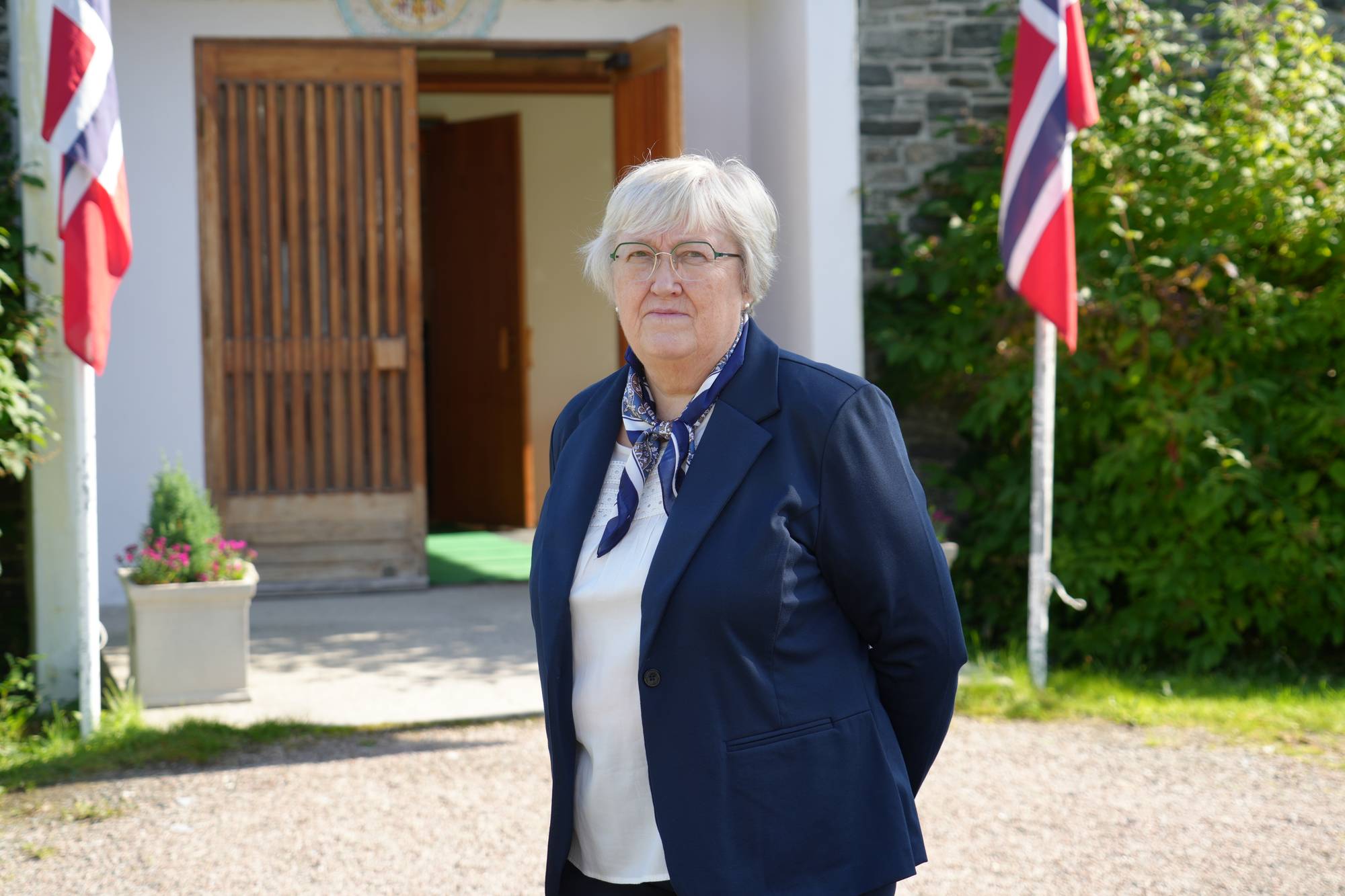 Kvinne med briller og mørk jakke står foran en bygning med to norske flagg på hver side. (Bildebeskrivelsen er laget av en KI-tjeneste)