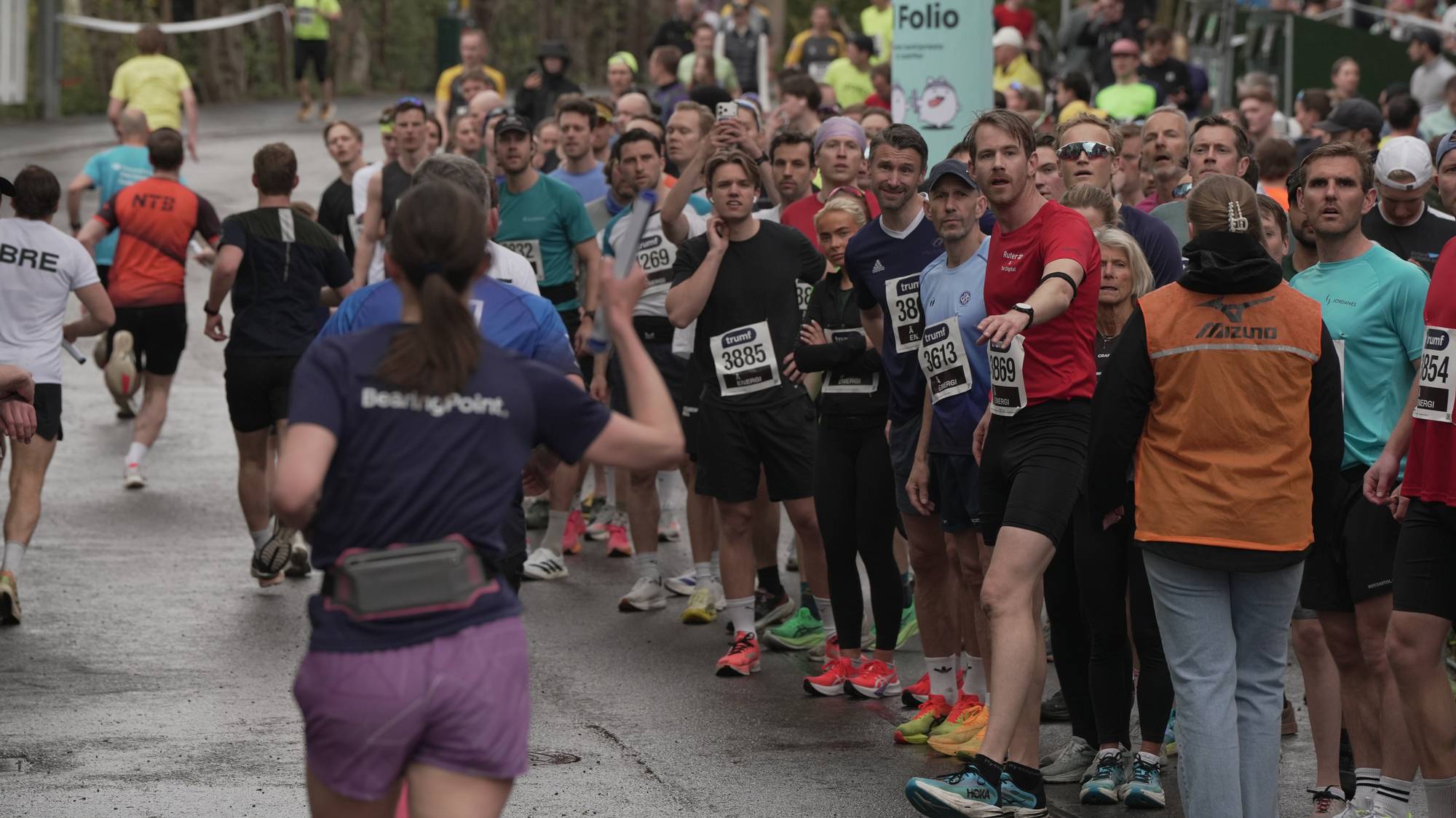 Et stort antall deltakere står klare ved startstreken i et løp. De fleste er kledd i sportsantrekk og har startnummer festet på brystet. I bakgrunnen ses tilskuere og et banner med en figur og tekst. En kvinne i en lilla shorts og t-skjorte er synlig i forgrunnen. Løypa er våt, noe som tyder på nylig regn. (Bildebeskrivelsen er laget av en KI-tjeneste)