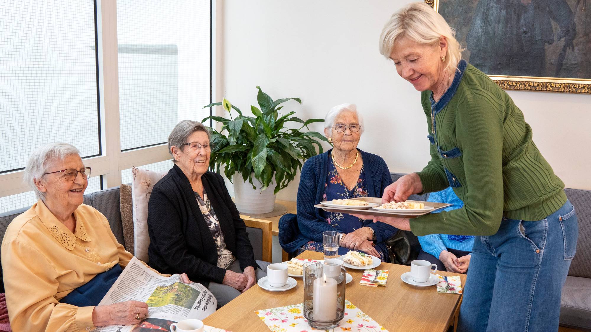 En kvinne serverer kaker til tre eldre kvinner som sitter rundt et bord i en stue. (Bildebeskrivelsen er laget av en KI-tjeneste)