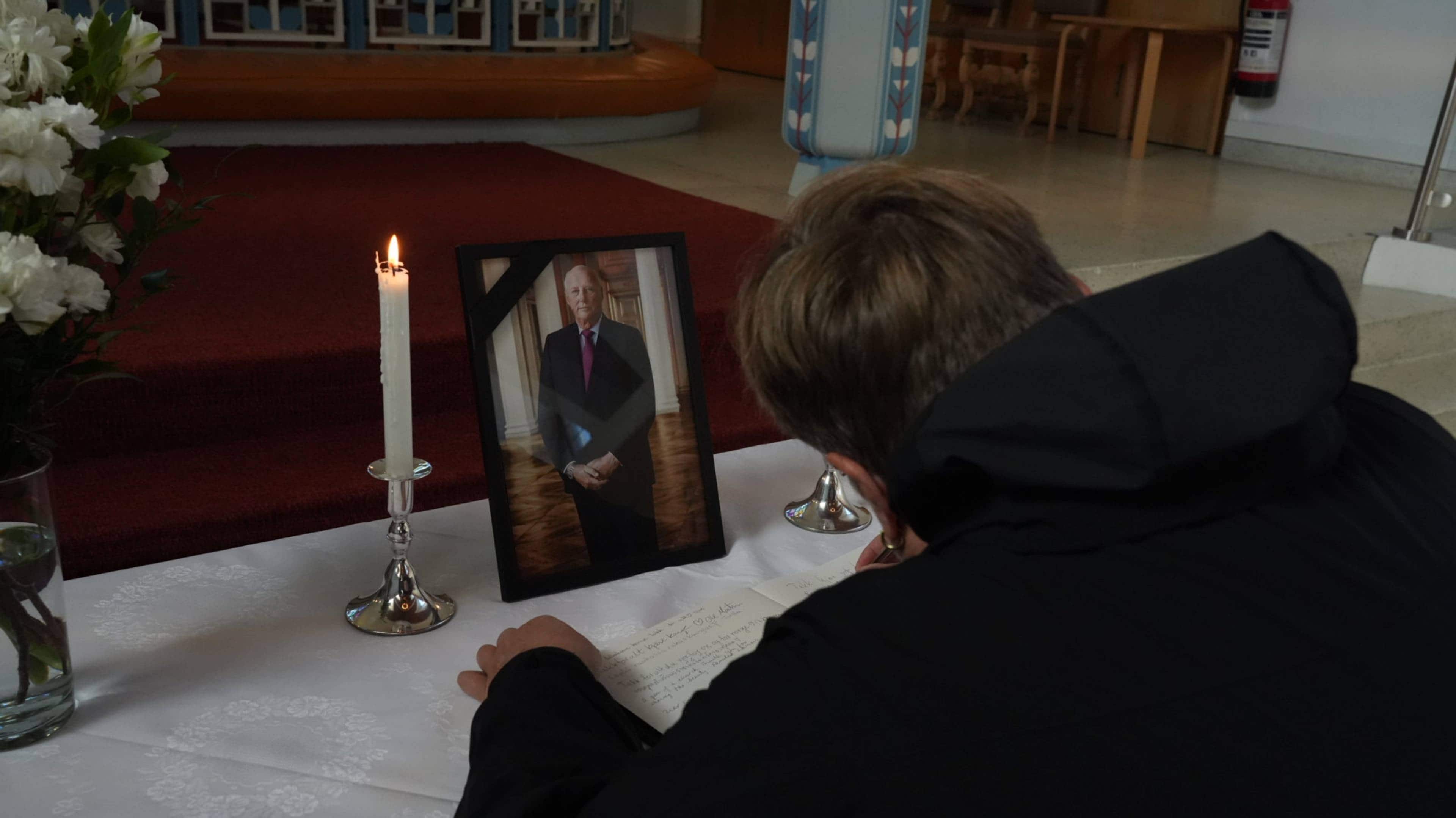 En person skriver i en kondolanseprotokoll ved siden av et bilde av kong Harald. (Bildebeskrivelsen er laget av en KI-tjeneste)