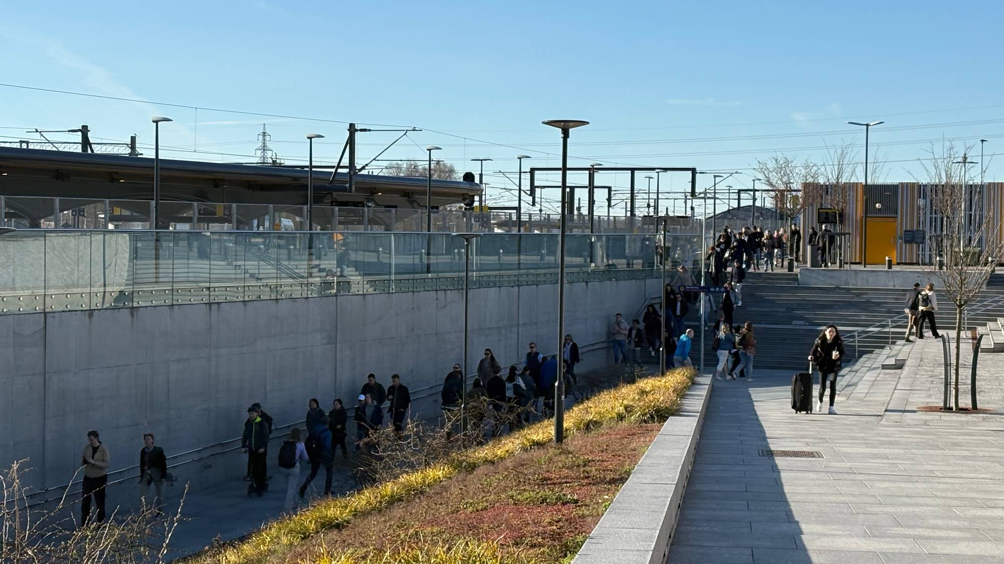 Folk går på en utendørs trapp ved en togstasjon. Flere personer med bagasje beveger seg oppover i bildet. En klar himmel dekker den øvre delen av motivet. (Bildebeskrivelsen er laget av en KI-tjeneste)