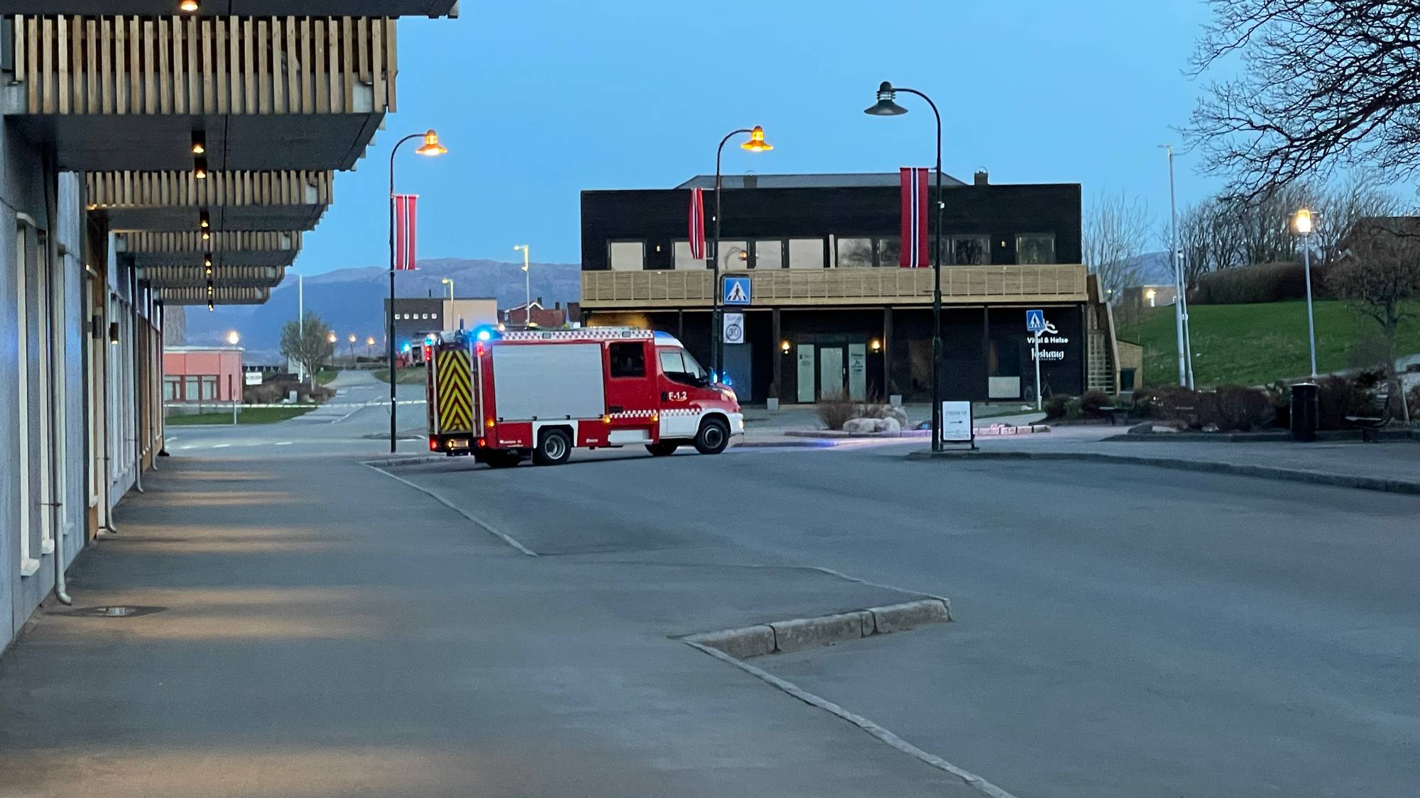 Brannbil med påslått blålys står parkert ved en bygning. Landskapet viser et åpent område med fjell i det fjerne. Bildet er tatt utendørs i skumring. (Bildebeskrivelsen er laget av en KI-tjeneste)