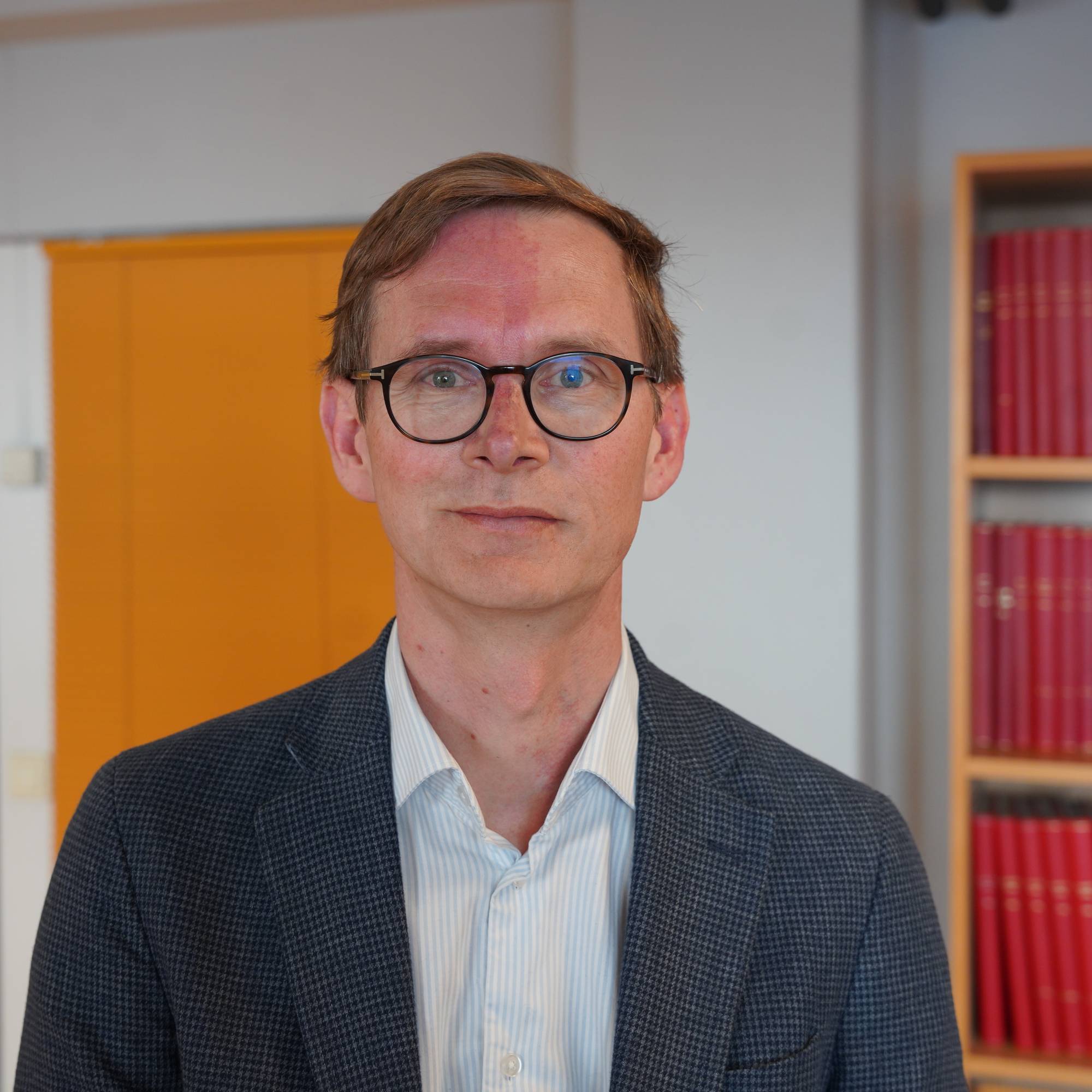En mann med briller iført skjorte og blazer ser rett inn i kamera. Han har sideskill, og et lite smil om munnen