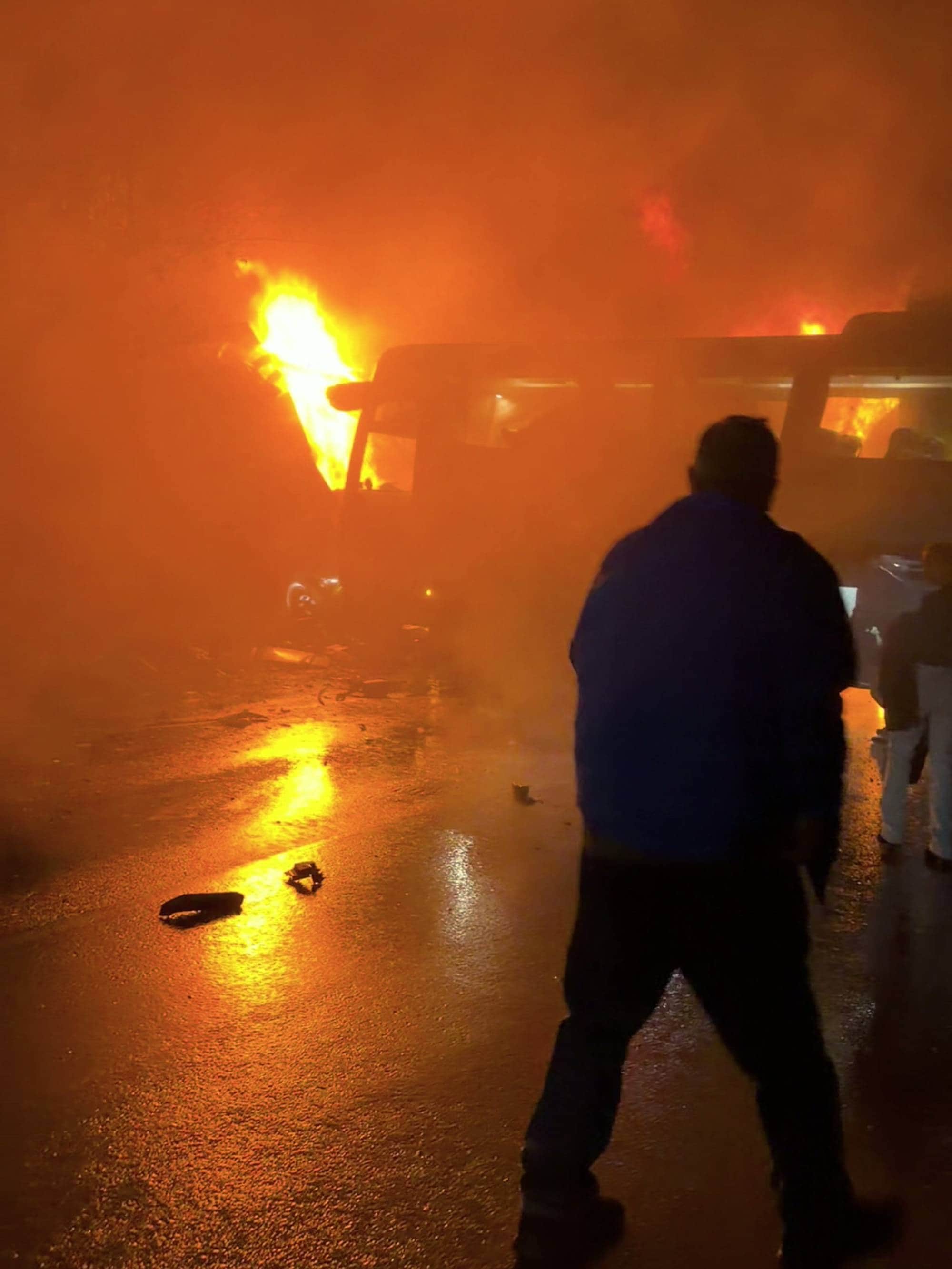En person står foran en brennende buss på en våt vei om natten. Store flammer lyser opp området. (Bildebeskrivelsen er laget av en KI-tjeneste)
