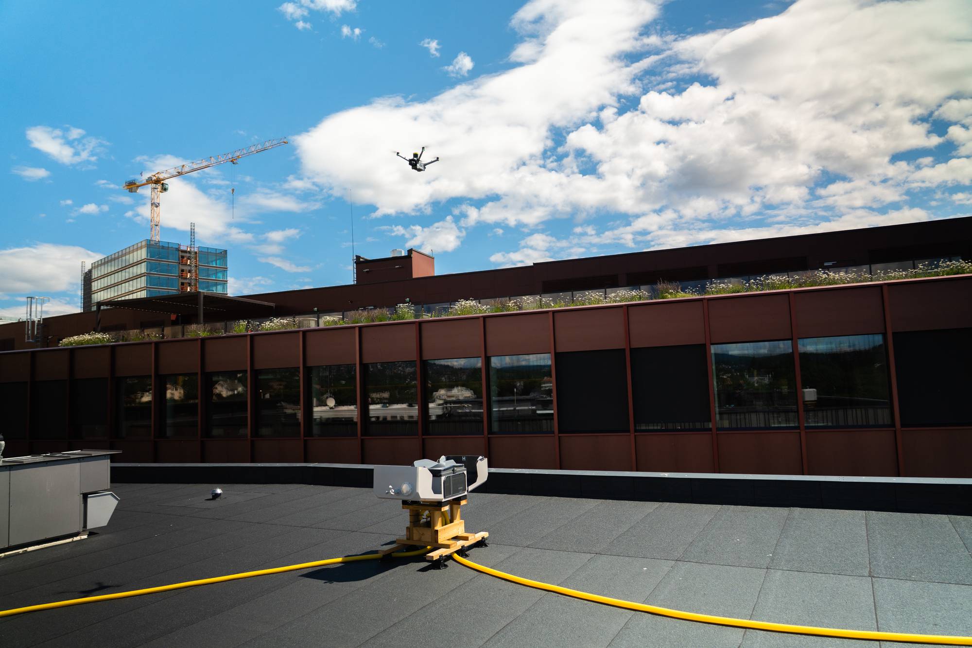 En drone flyr over en bygning med et grønt tak. Det pågår byggearbeid med en kran i bakgrunnen. Solen skinner fra en blå himmel med hvite skyer. På taket er det et apparat og en gul slange. (Bildebeskrivelsen er laget av en KI-tjeneste)
