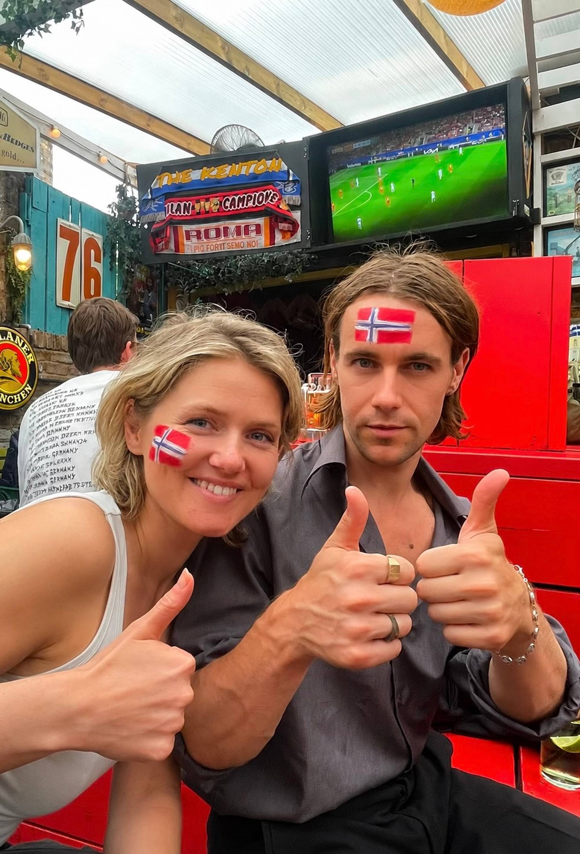 En mann og en kvinne som sitter sammen på en pub. Bak dem henger det en TV-skjerm som viser en fotballkamp. De har det norske flagget malt i ansiktet og poserer smilende med tommelen opp.