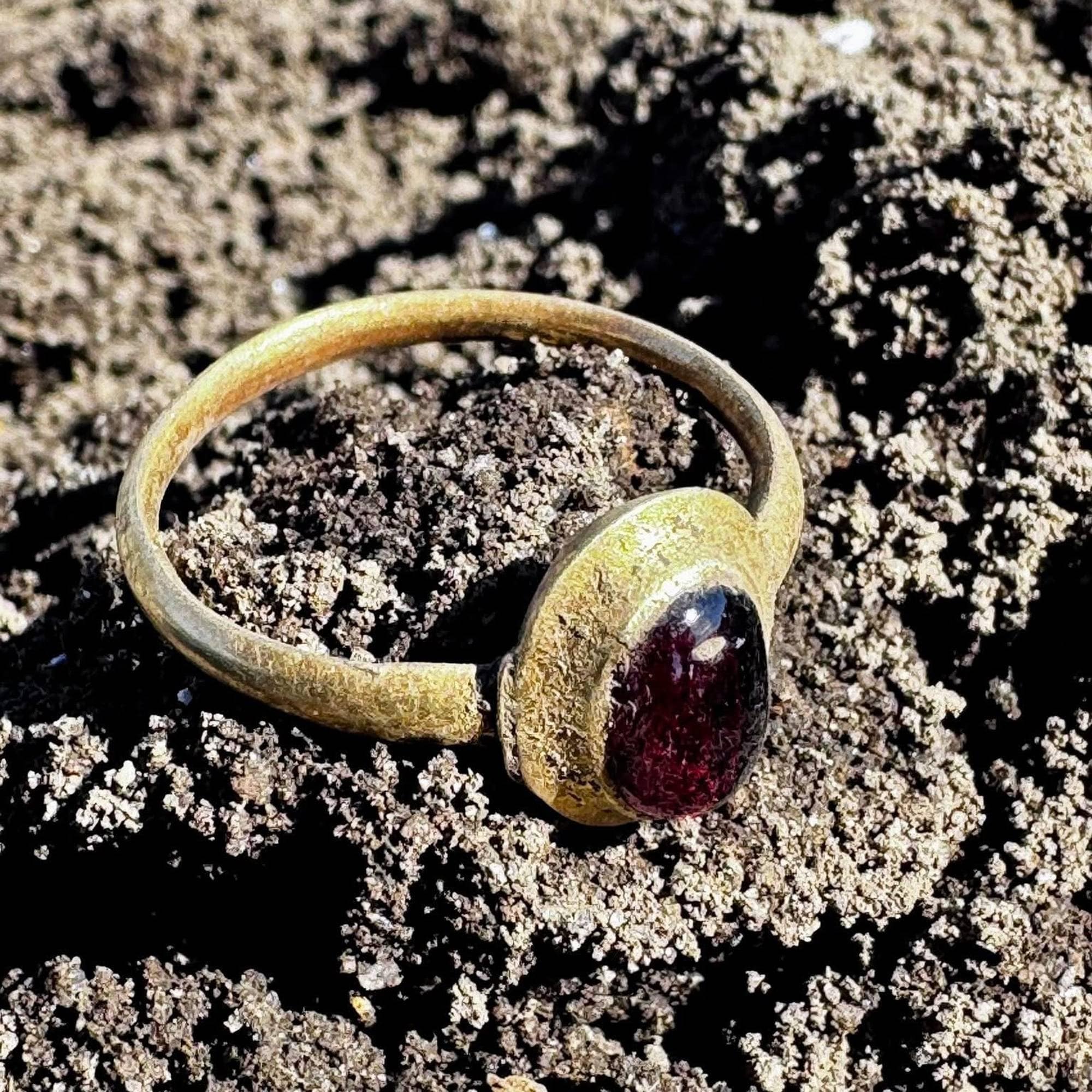 En gullring med en mørkerød stein ligger på bakken. Ringen har en enkel utforming med en dekorativ innfatning. Bakken består av jord og sand. (Bildebeskrivelsen er laget av en KI-tjeneste)