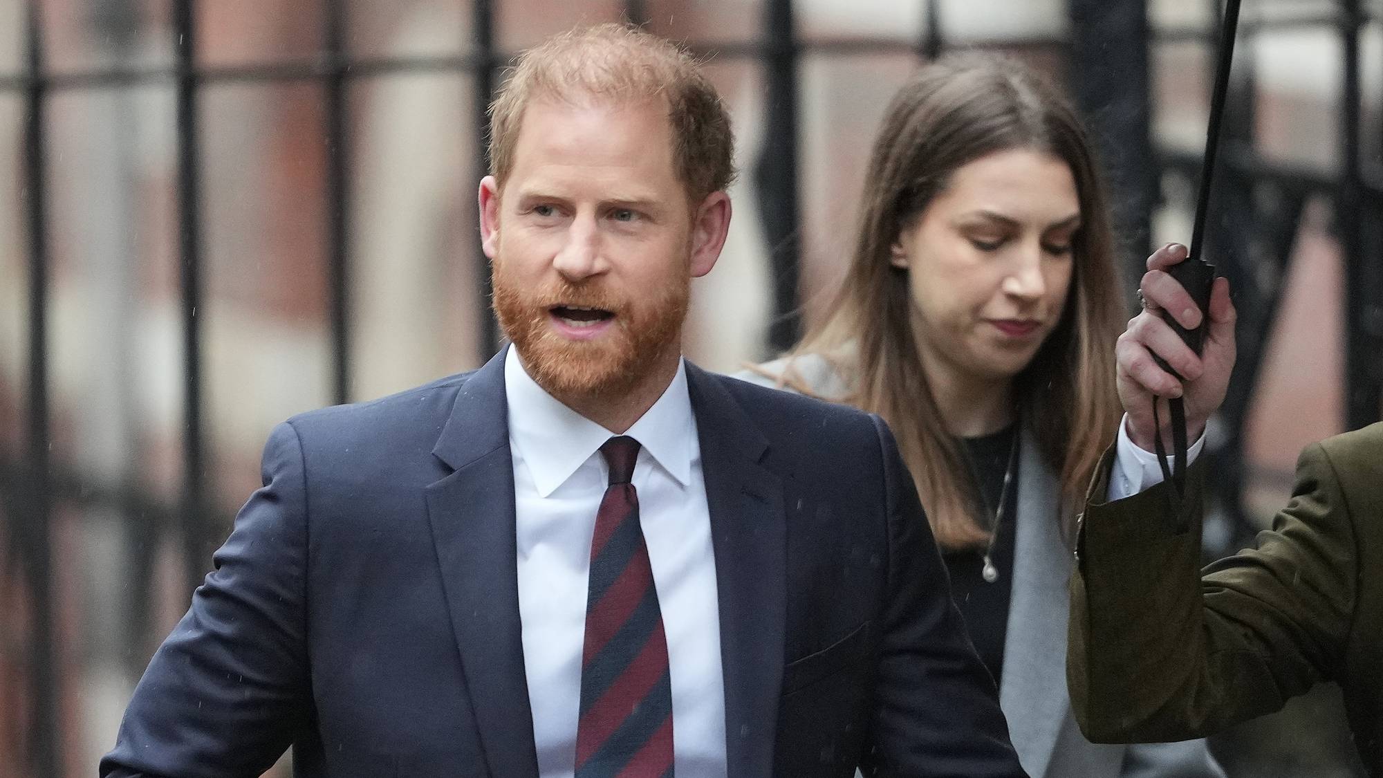 Prins Harry i mørk dress og slips foran et gjerde. En kvinne med paraply går ved siden av ham. (Bildebeskrivelsen er laget av en KI-tjeneste)