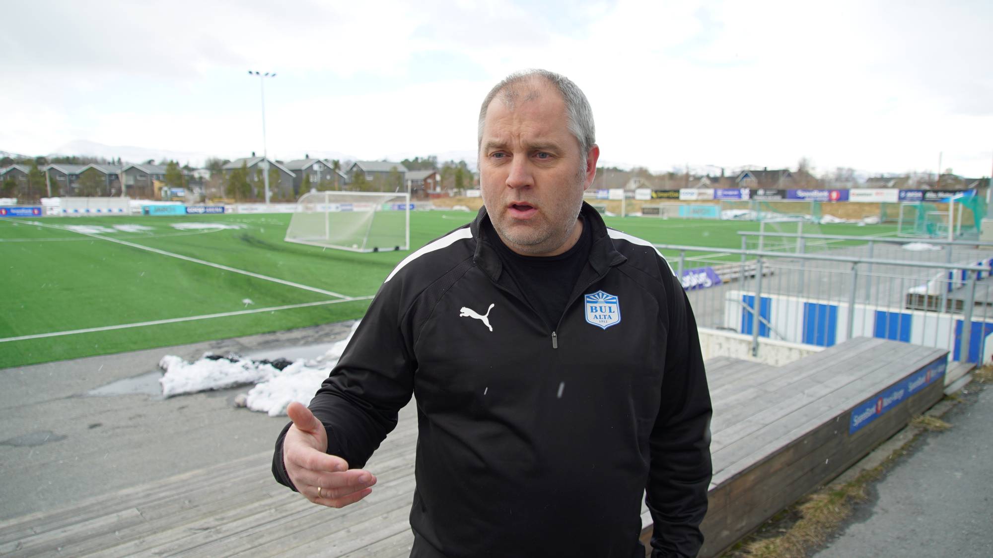 En mann med sort genser ved en utendørs fotballbane.