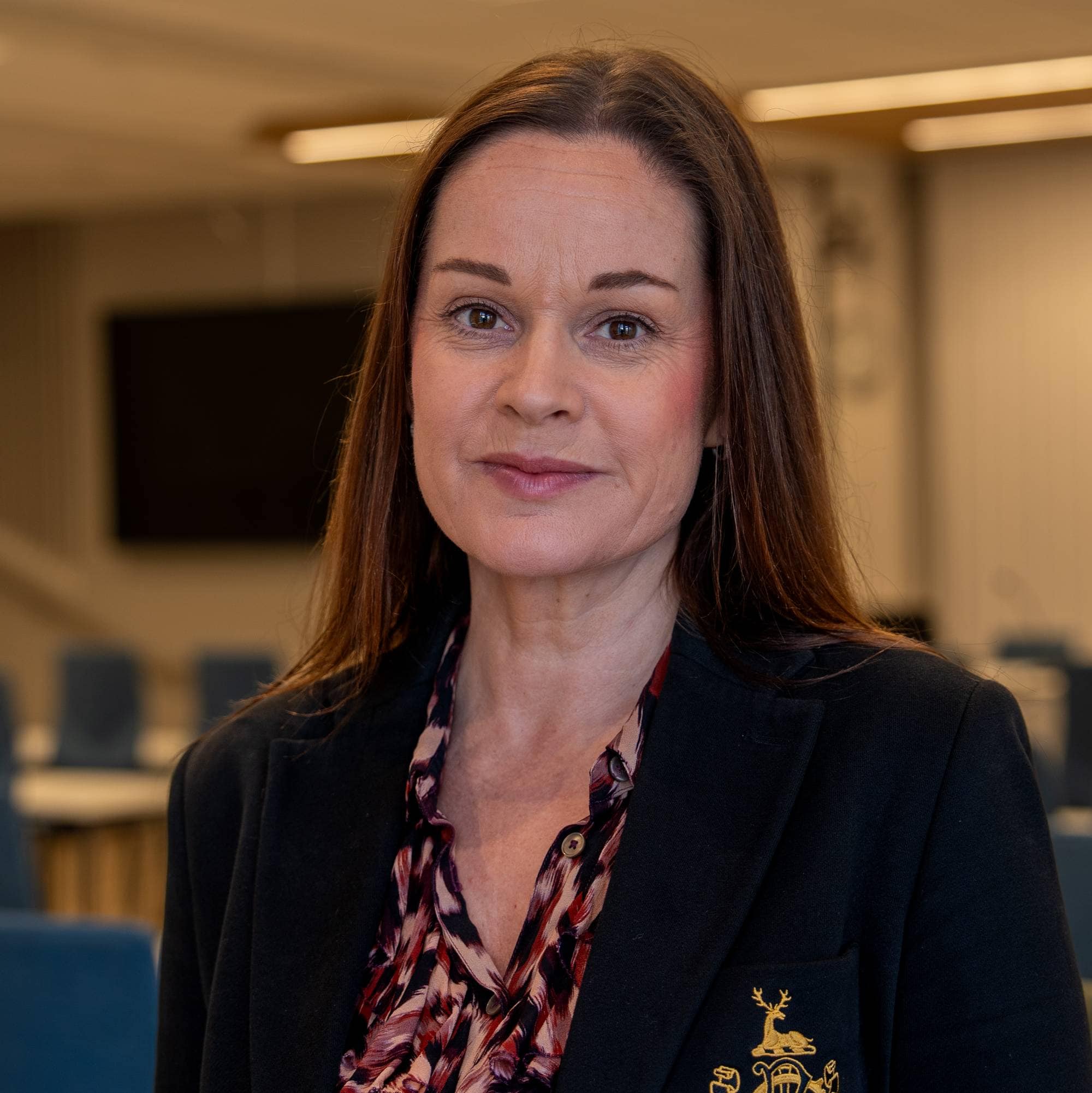 Portrett av kvinne i en mørk blazer foran tomme stoler i et møterom. (Bildebeskrivelsen er laget av en KI-tjeneste)