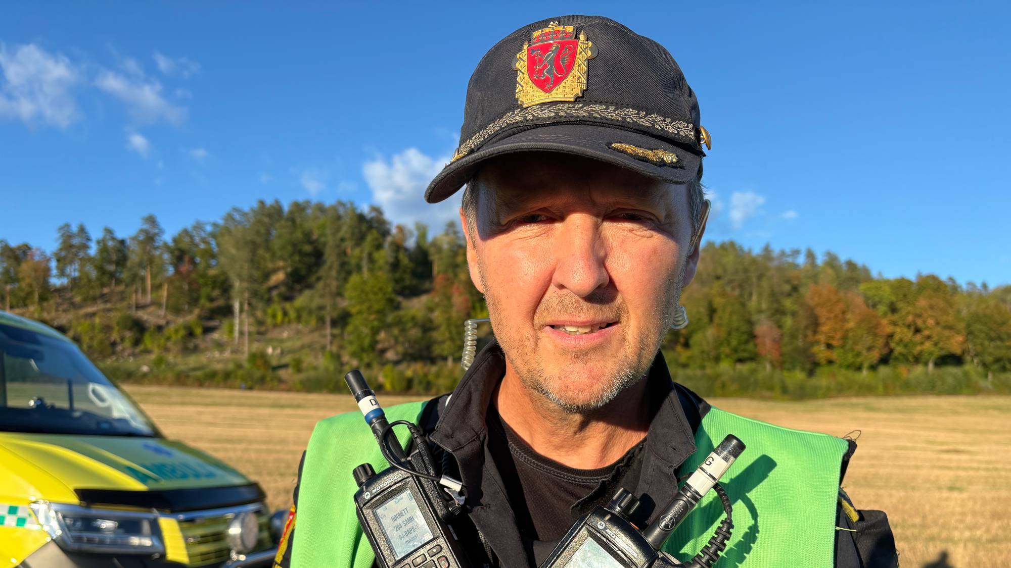 Politimann med refleksvekst og mobiltelefoner står ved en politibil.