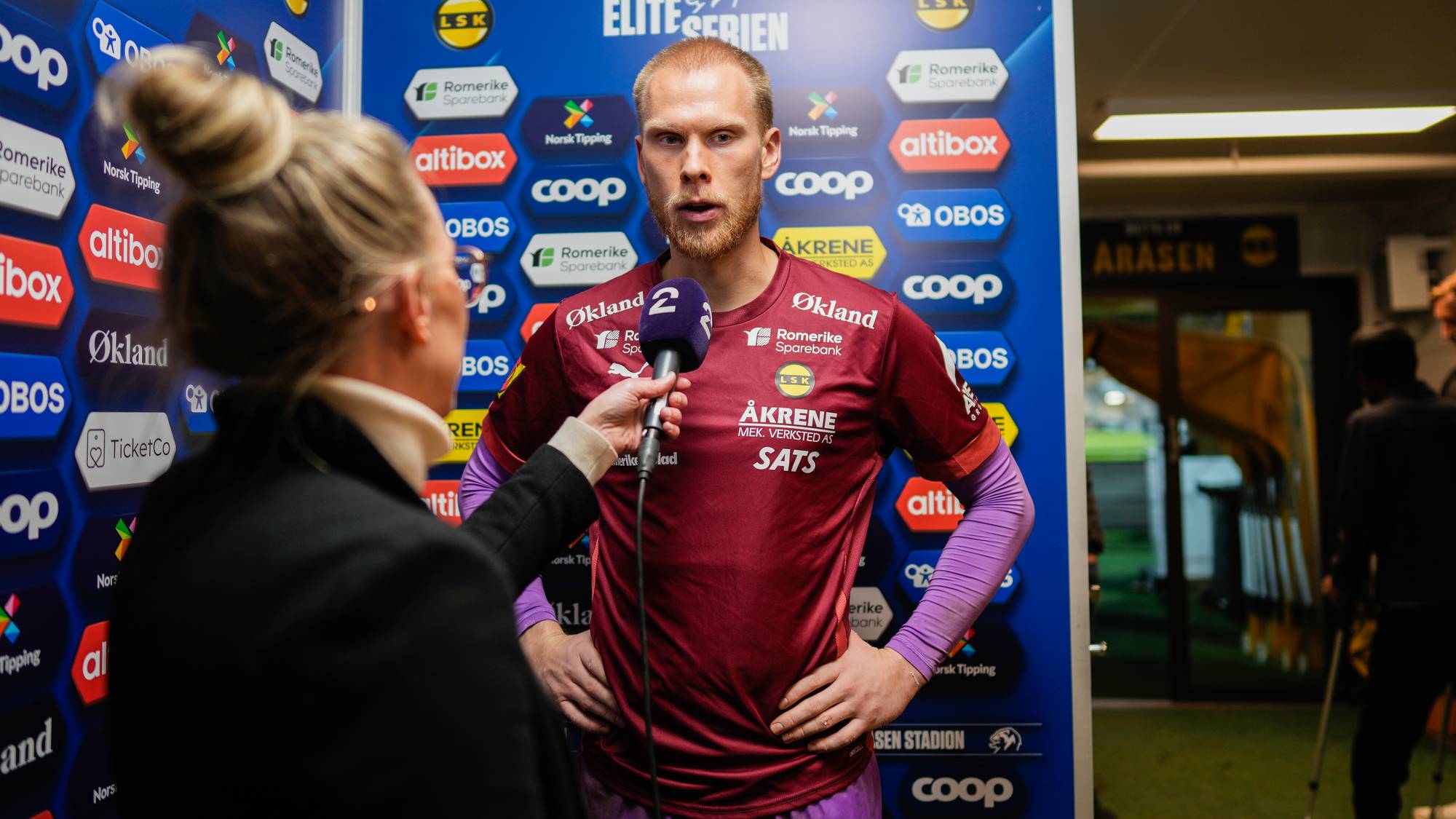 En fotballspiller blir intervjuet foran en vegg med sponsorer.
Han bærer en rød og lilla drakt med nummer 2.
Kvinnen som intervjuer ham har en svart dressjakke.
Bakgrunnen viser ulike logoer. (Bildebeskrivelsen er laget av en KI-tjeneste)