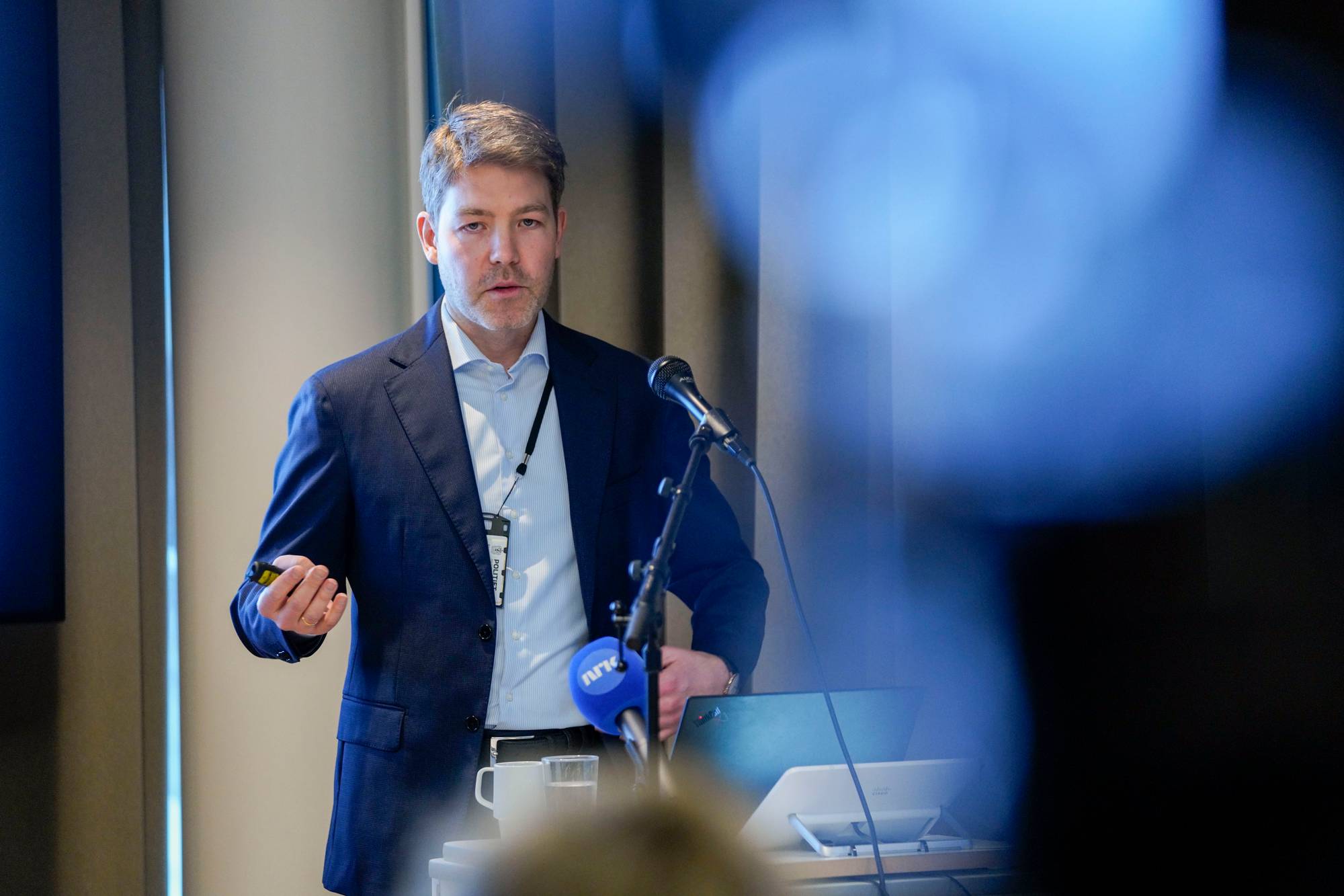 Mann i dress holder presentasjon foran mikrofoner. (Bildebeskrivelsen er laget av en KI-tjeneste)