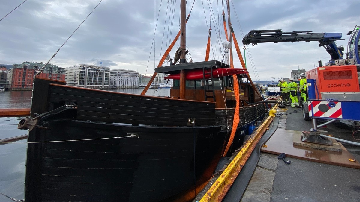 Arbeidere i gule vester bistår med å løfte en mørk trebåt med kran. Båten er festet med oransje stropper. I bakgrunnen ses flere bygningsmasser og en overskyet himmel. (Bildebeskrivelsen er laget av en KI-tjeneste) FOTO: Sandra Amalie Lid Krumsvik/NRK