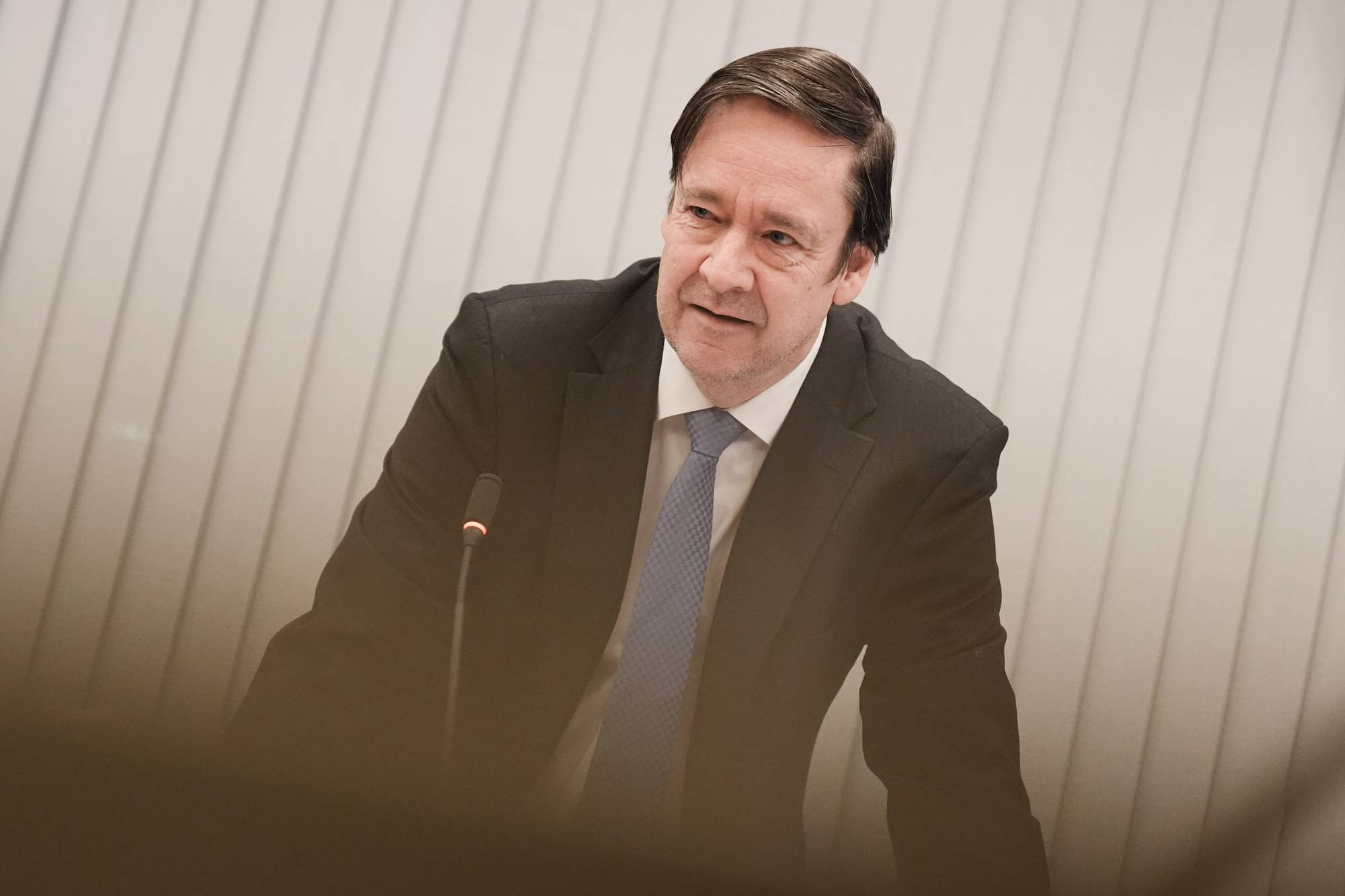 En mann iført mørk dress og blått slips står ved et podium. Han er i ferd med å holde en presentasjon, med mikrofon plassert foran seg. Omgivelsene består av en lys bakgrunn med horisontale striper, som gir en formell setting. Stemningen kan tolkes som alvorlig, gitt konteksten av en presentasjon. (Bildebeskrivelsen er laget av en KI-tjeneste)