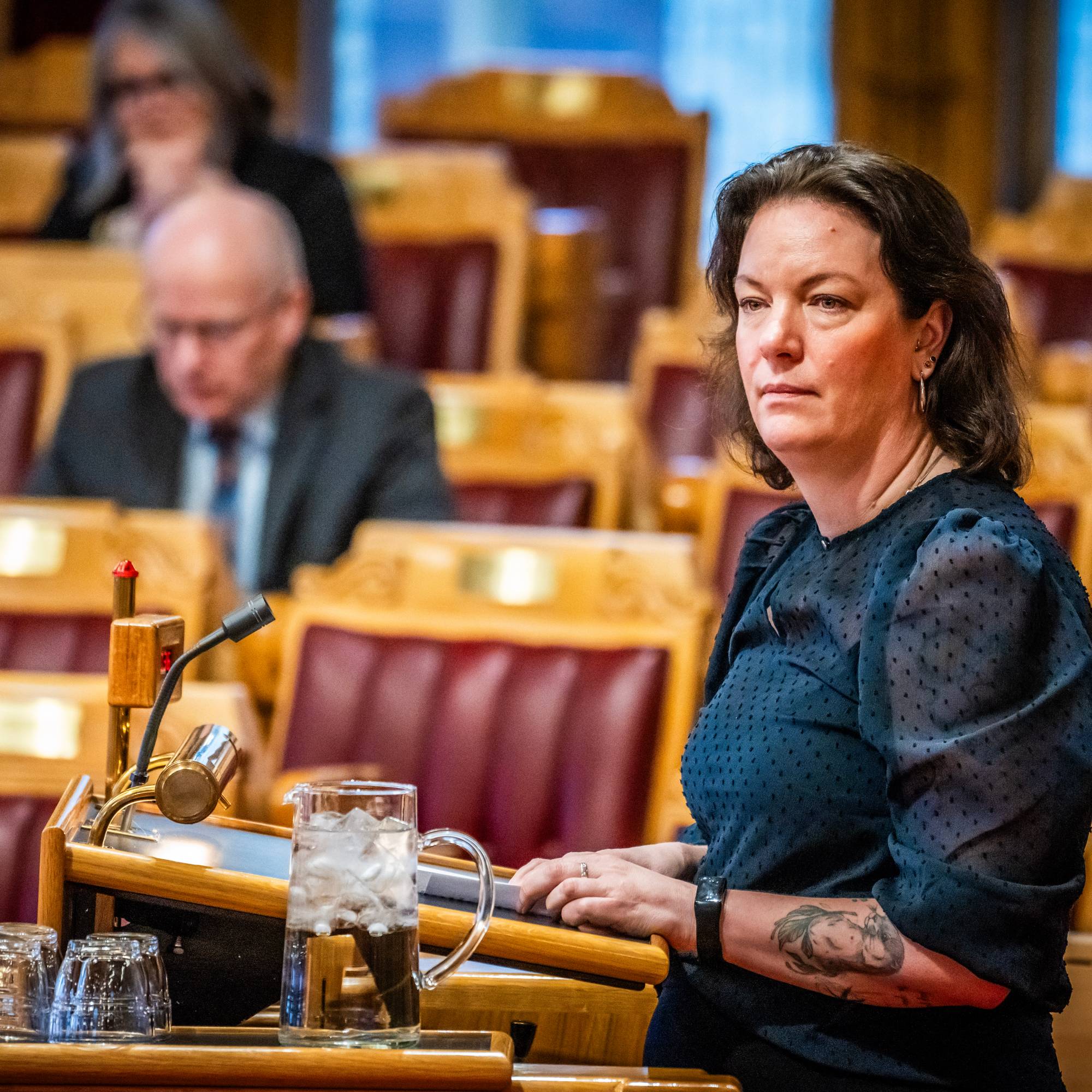 En kvinne sitter foran en talerstol med et glass vann og to tomme glass. Hun er i fokus i bildet, mens andre personer sitter lenger bak i en sal med mange seter. Det er et NTB-merke synlig i bildet. (Bildebeskrivelsen er laget av en KI-tjeneste)