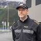 Marius André Jenssen Stenberg / NRK En mann i politiuniform står foran en bygning og et fjellandskap. Uniformen er svart med gule skulderklaffer og et rutete bånd over brystet. Mannen har på seg en caps med et riksvåpen. I bakgrunnen er det et veikryss med biler og flagg. (Bildebeskrivelsen er laget av en KI-tjeneste)