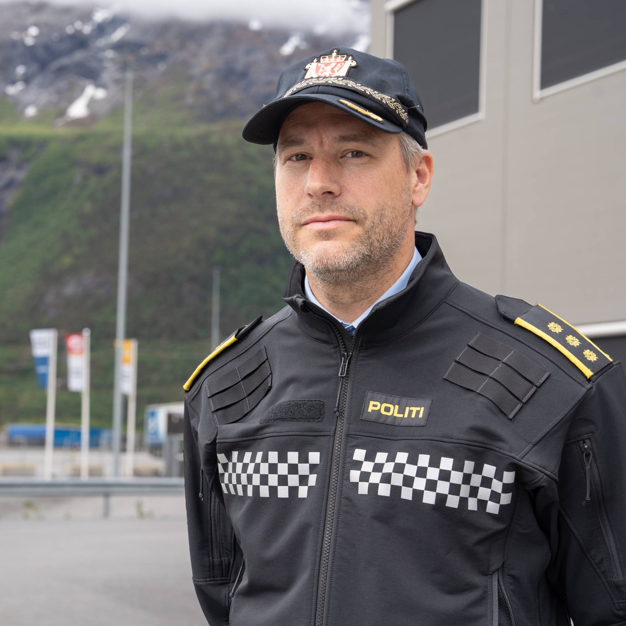 En mann i politiuniform står foran en bygning og et fjellandskap. Uniformen er svart med gule skulderklaffer og et rutete bånd over brystet. Mannen har på seg en caps med et riksvåpen. I bakgrunnen er det et veikryss med biler og flagg. (Bildebeskrivelsen er laget av en KI-tjeneste)