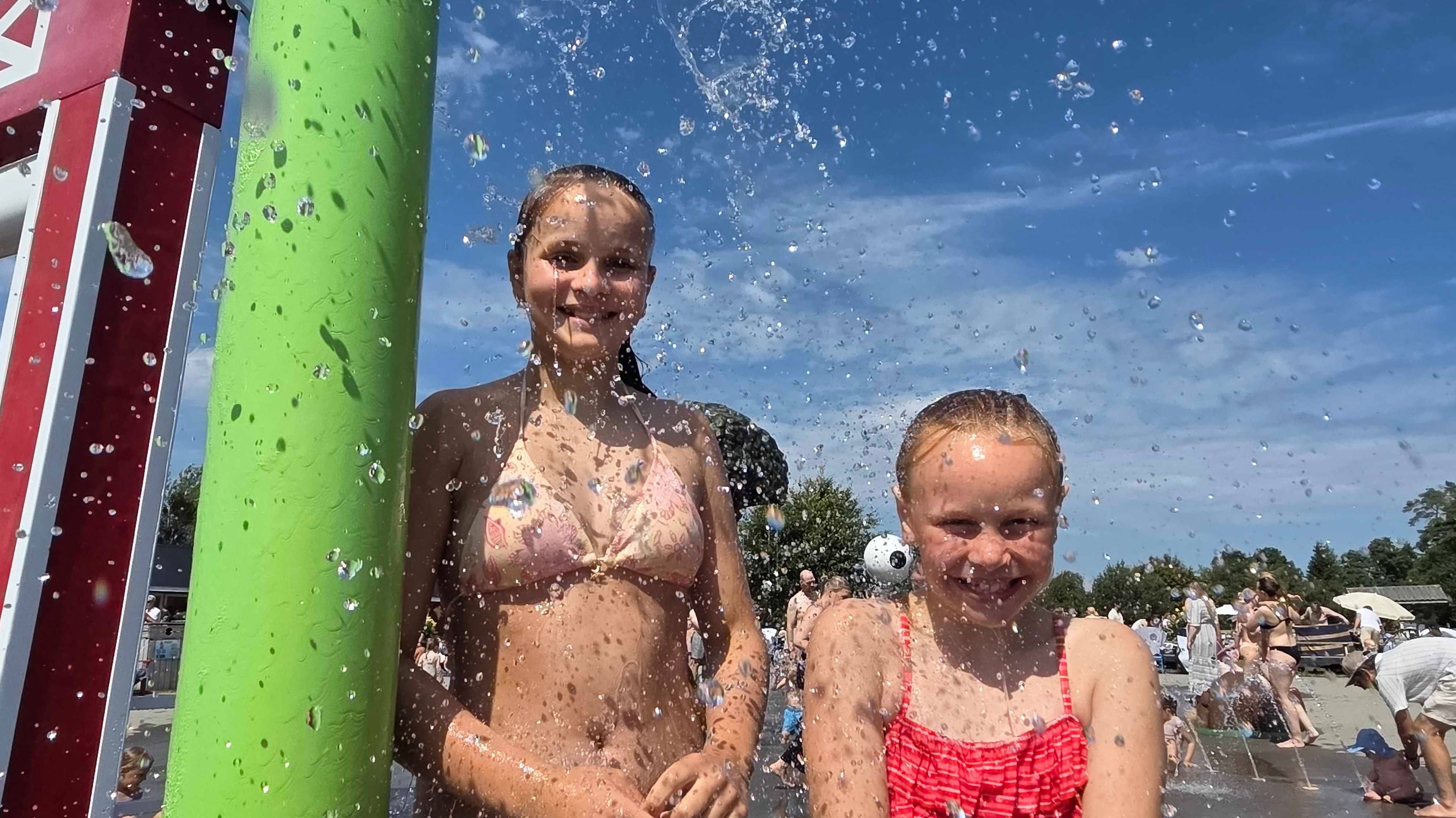 To yngre jenter står under vannspruten og nyter finværet i familieparken.