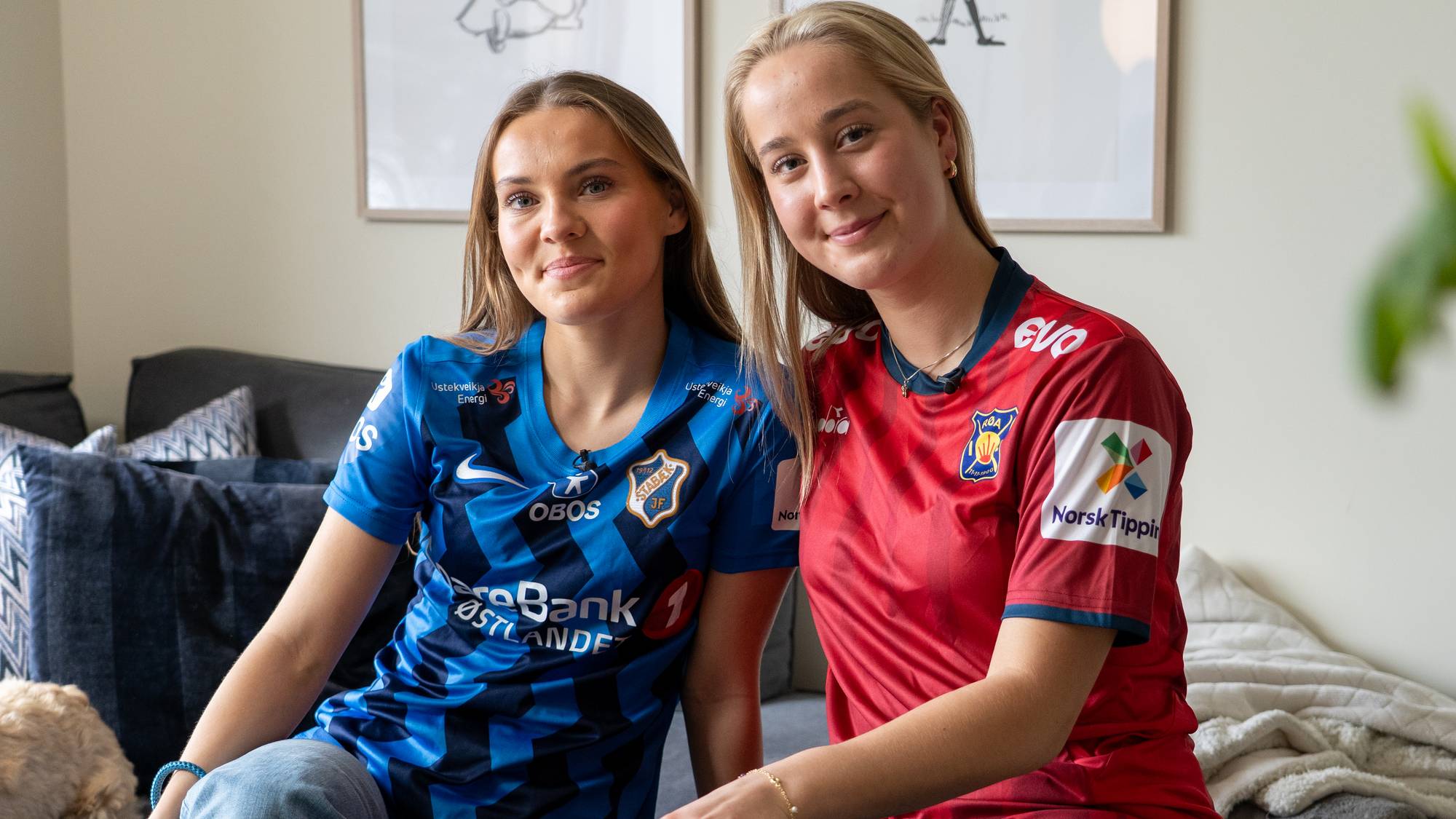 To unge kvinner sitter på en sofa og ser mot kamera. Kvinnen til venstre har på seg en blå fotballtrøye, mens kvinnen til høyre har på seg en rød fotballtrøye. De er i et rom med bilder på veggen og puter i sofaen. (Bildebeskrivelsen er laget av en KI-tjeneste)