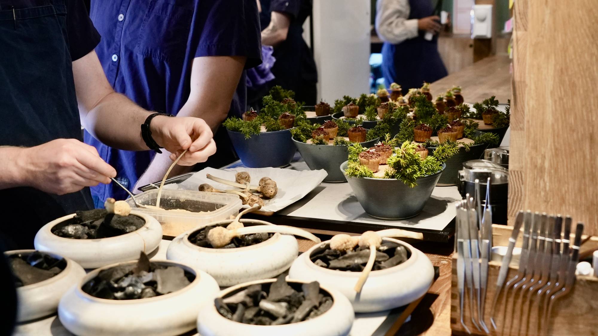 Personer forbereder mat i en restaurant. Det er flere små retter med grønt og kjøtt i skåler. Foran ligger kull og ingredienser klare til bruk. (Bildebeskrivelsen er laget av en KI-tjeneste)