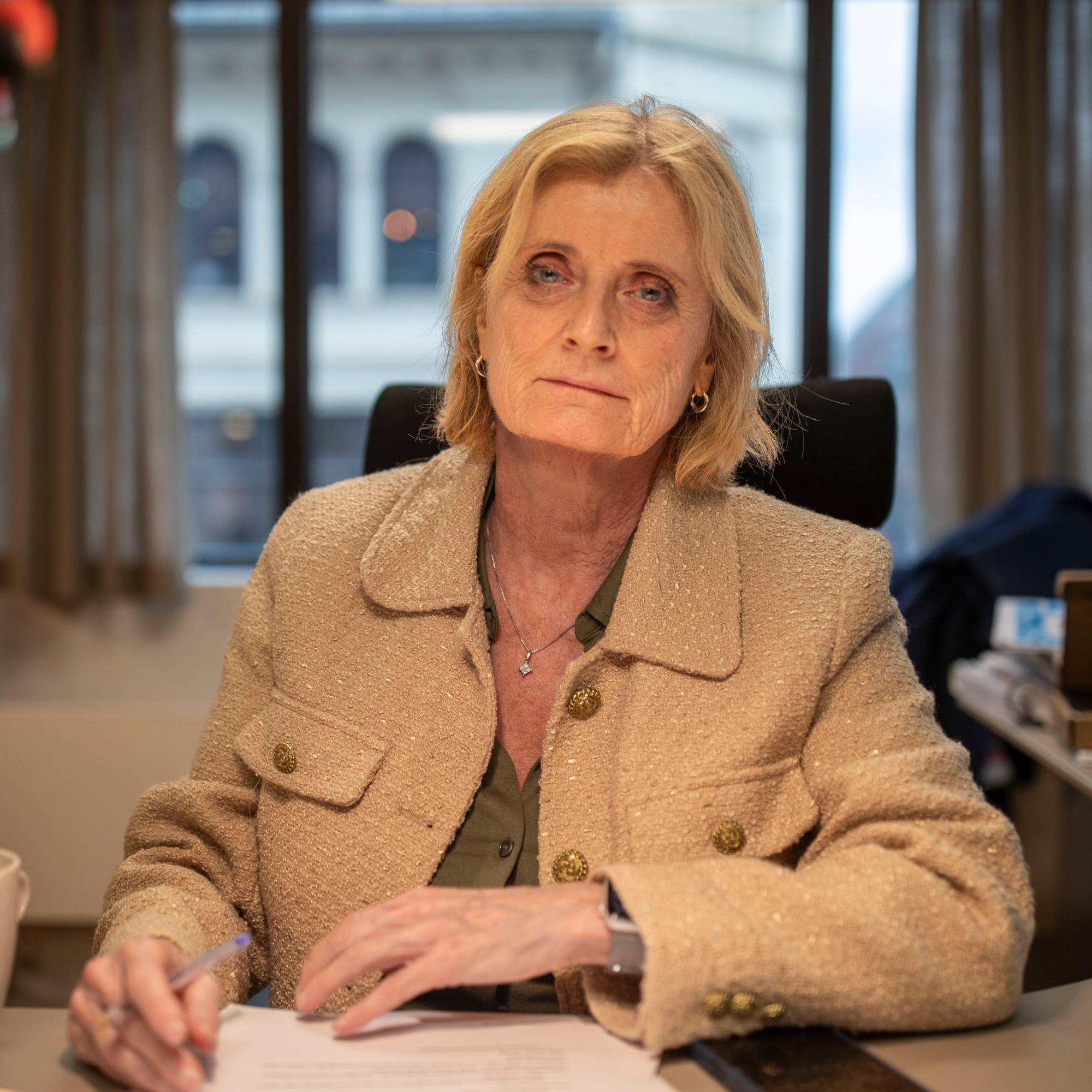 En kvinne med halvlagt, lyst hår ser inn i kamera. Hun har på seg en grønn bluse og en lys jakke.Hun sitter med et skrivebord med en penn i hånden.