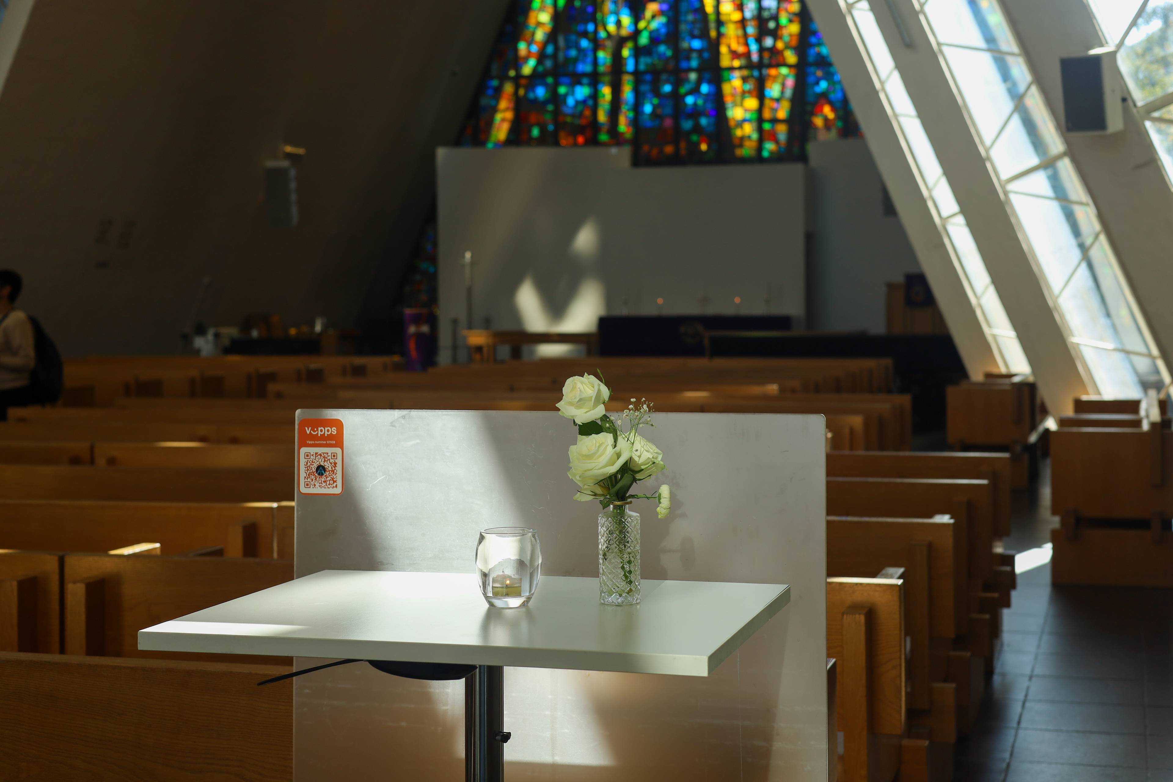 Hvite roser og et telys står på et bord foran kirkebenker i Ishavskatedralen. (Bildebeskrivelsen er laget av en KI-tjeneste)