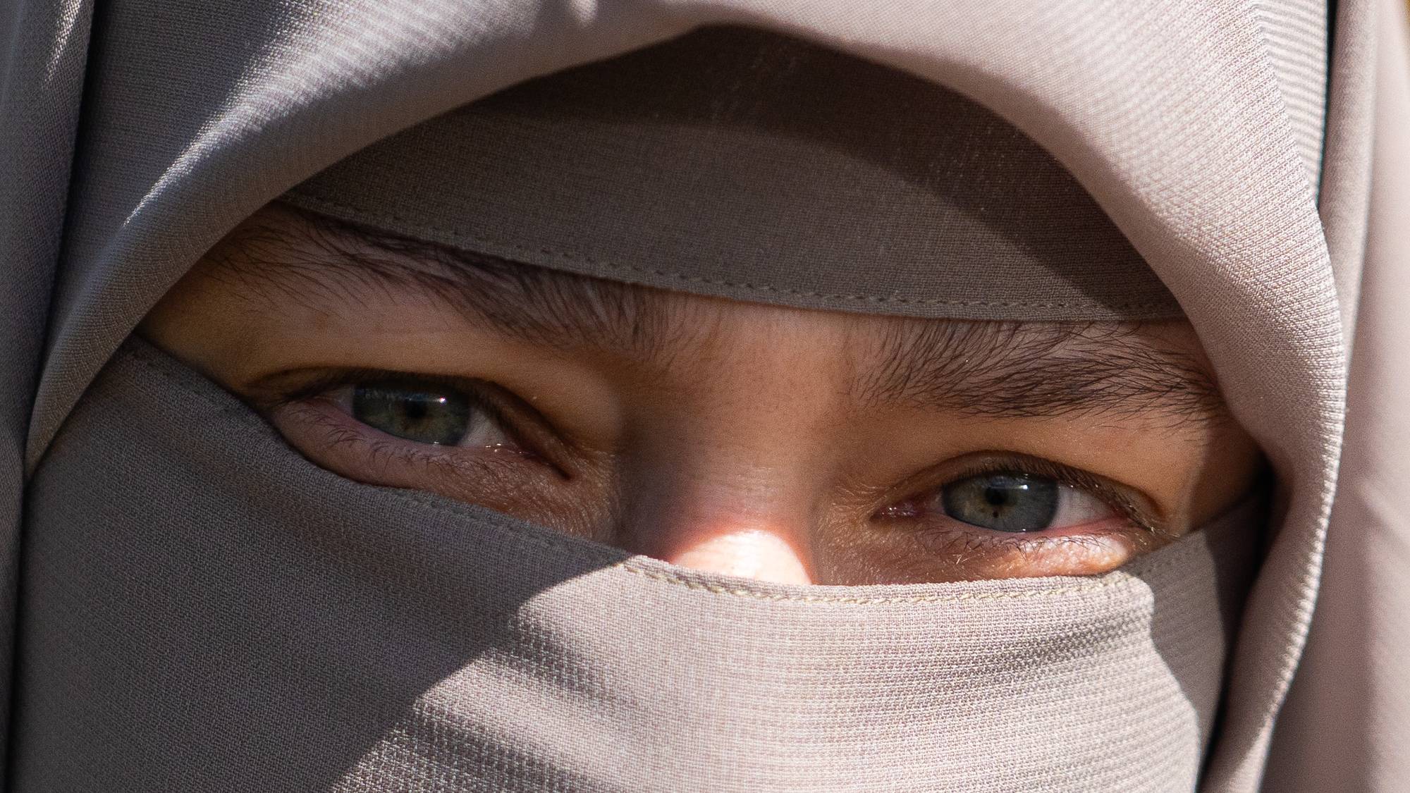 Et nærbilde av en person iført niqab. Personen har grå kledning og slør som dekker ansiktet, bortsett fra øynene. I bakgrunnen er det en uskarpt hus og grønt løvverk. (Bildebeskrivelsen er laget av en KI-tjeneste)