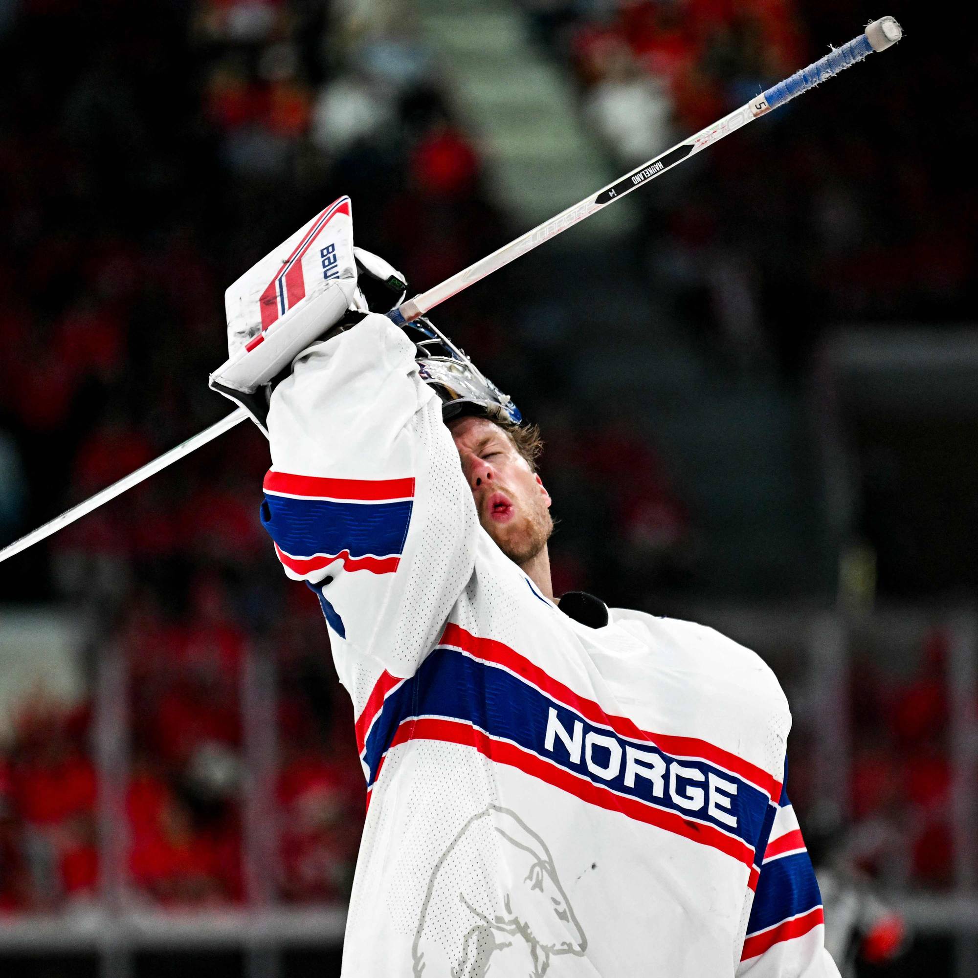 En ishockeymålvakt i Norges drakt jubler med køllen løftet. Publikum i bakgrunnen har røde drakter. Det er en ishockeykamp. (Bildebeskrivelsen er laget av en KI-tjeneste)