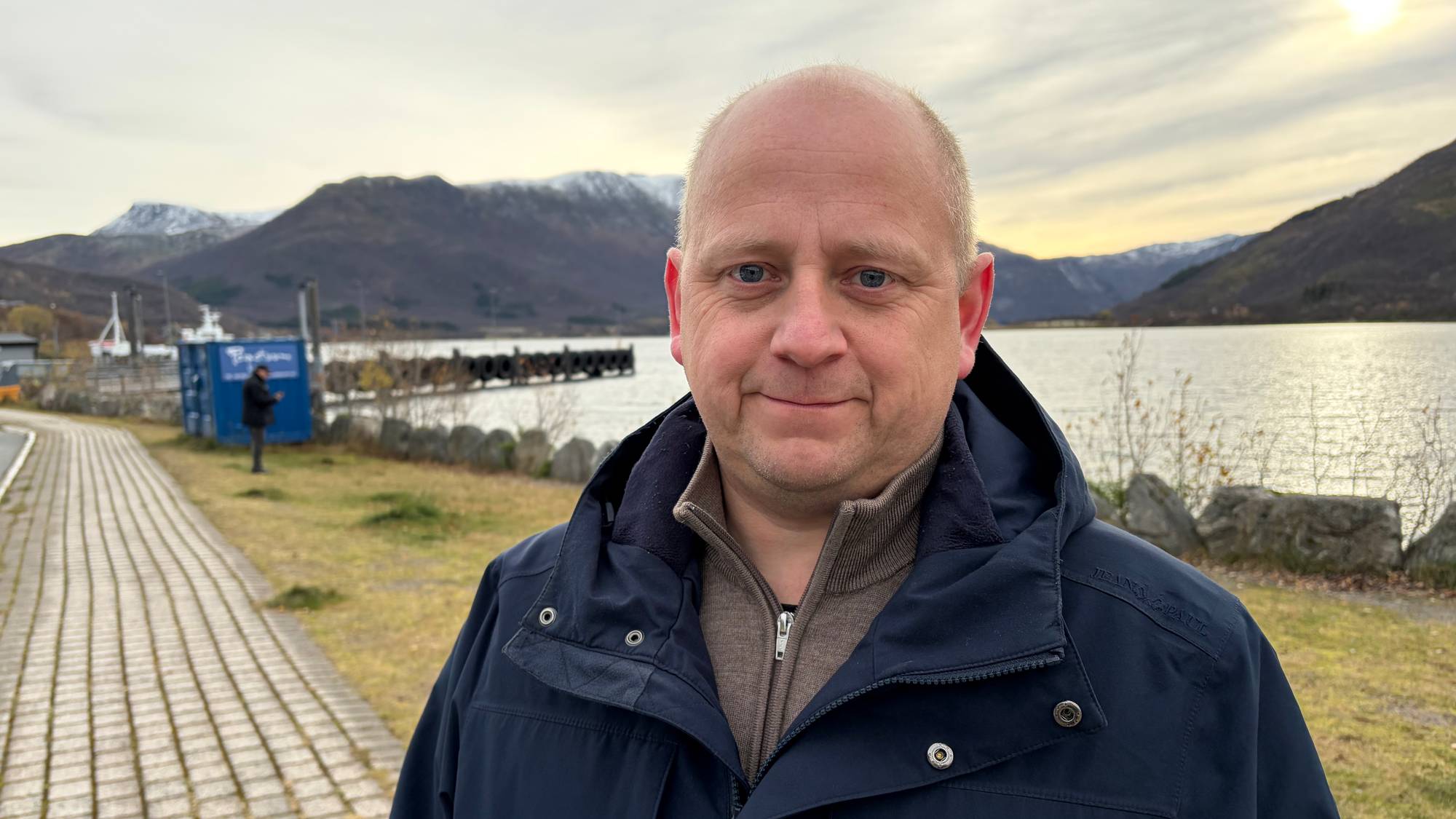 Mann i mørk jakke står ved en kai med fjell og havet i bakgrunnen. (Bildebeskrivelsen er laget av en KI-tjeneste)