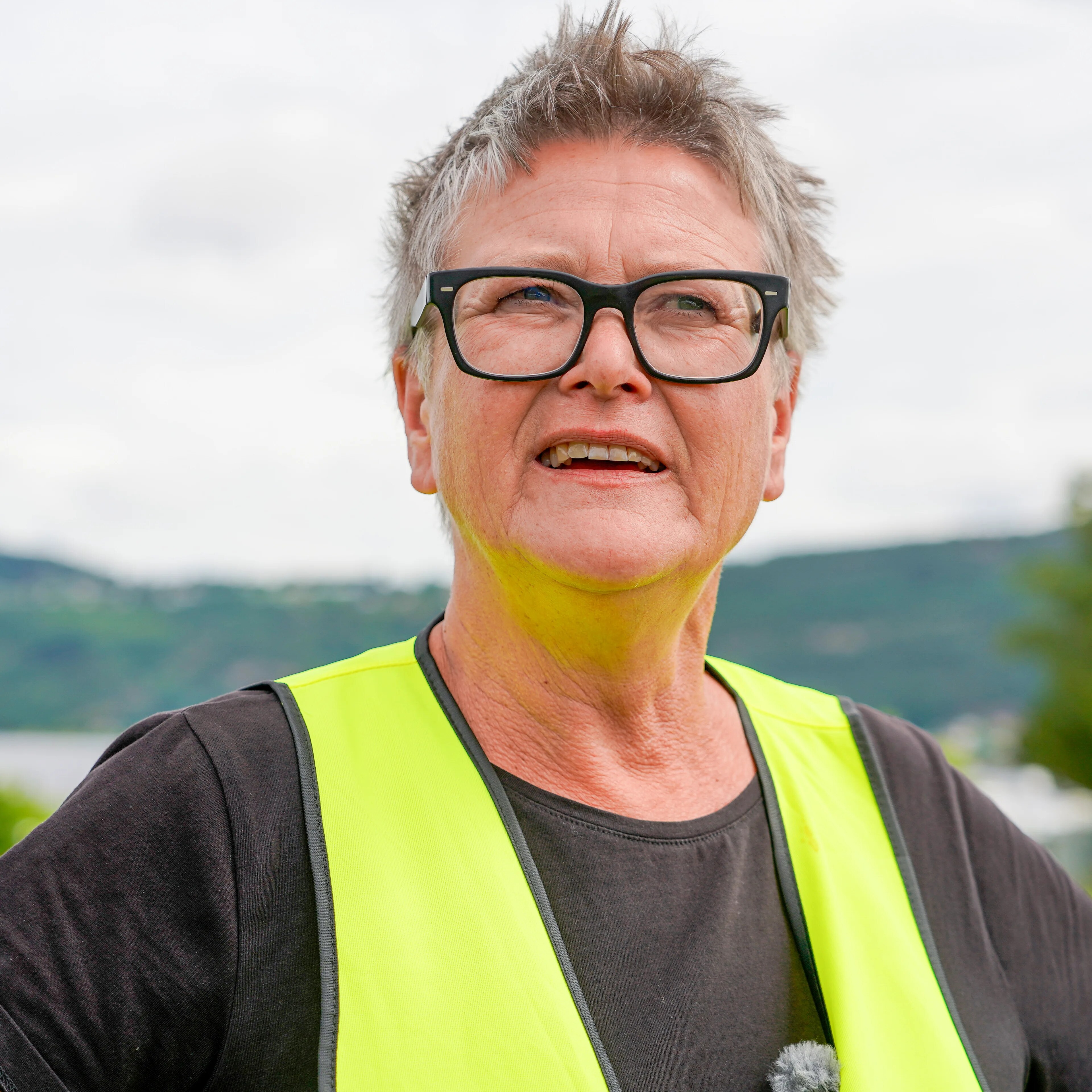 Ei kvinne med grått hår og briller står med henda på hofta og eit landskap i bakgrunnen
