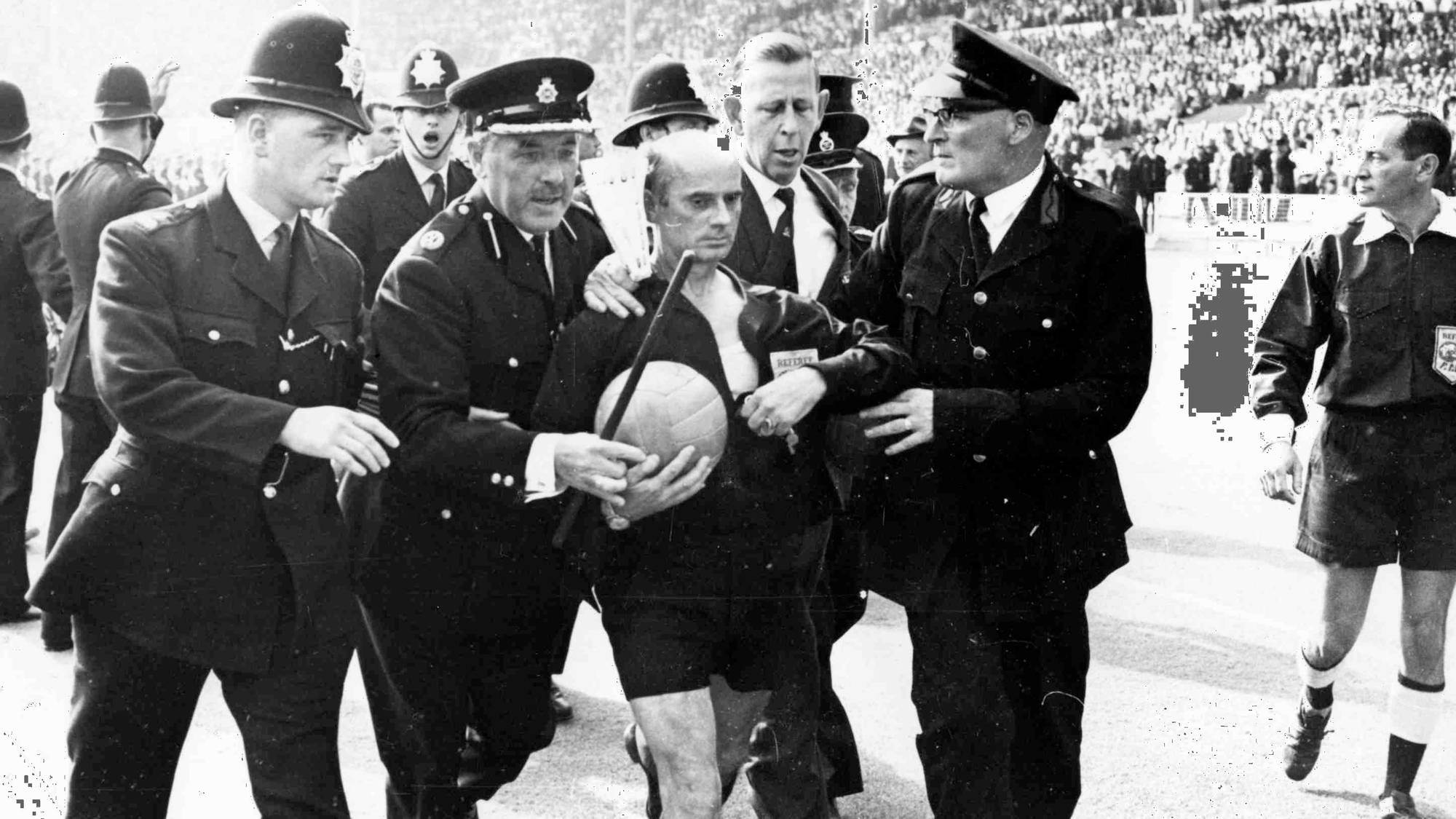 Politibetjenter eskorterer en fotballdommer av banen på et fullsatt stadion. Svart-hvitt fotografi. (Bildebeskrivelsen er laget av en KI-tjeneste)