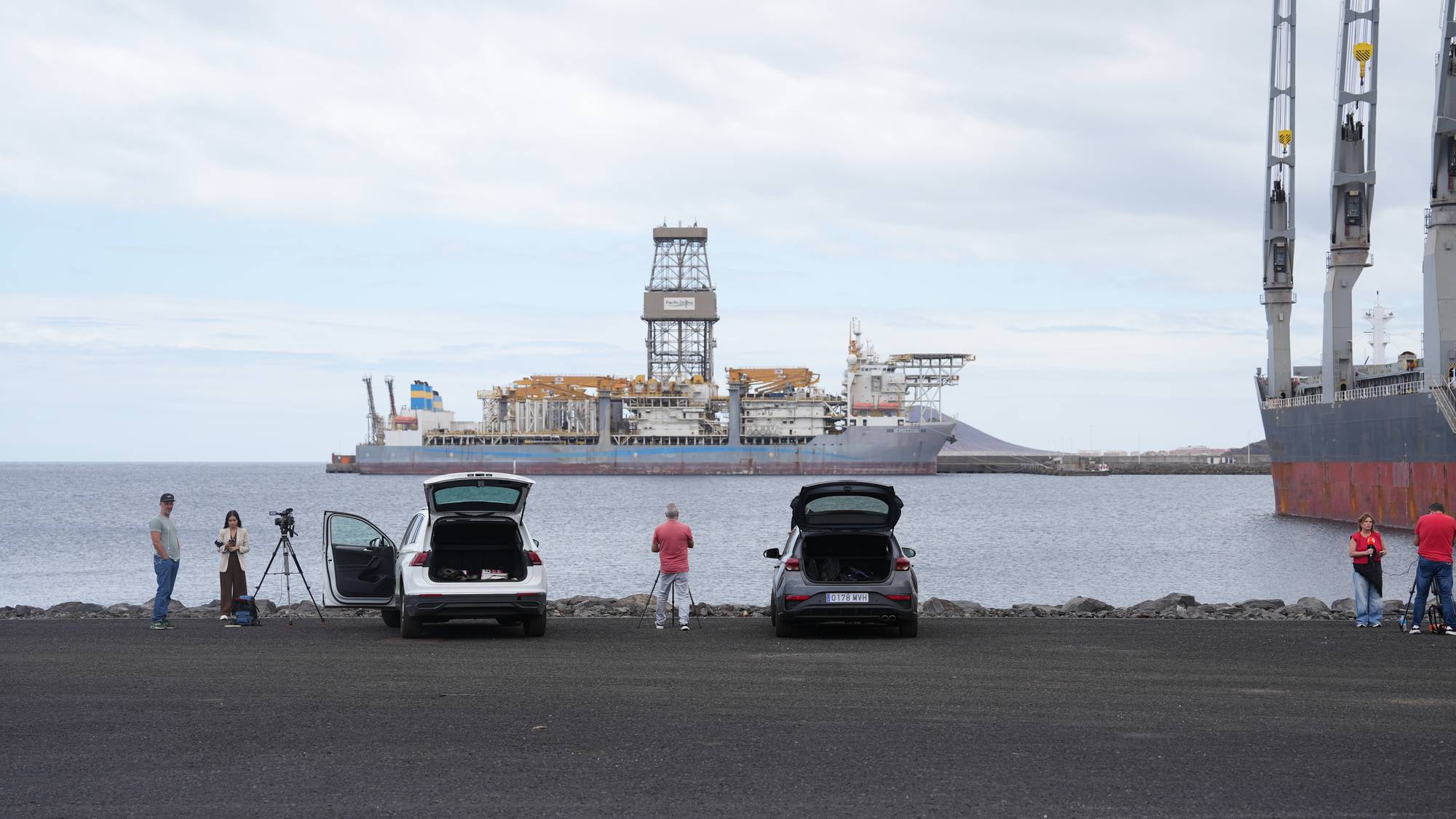 Et skip ligger til kai i en havn. To biler med åpne bagasjerom er parkert ved vannkanten. Personer står i nærheten av bilene.