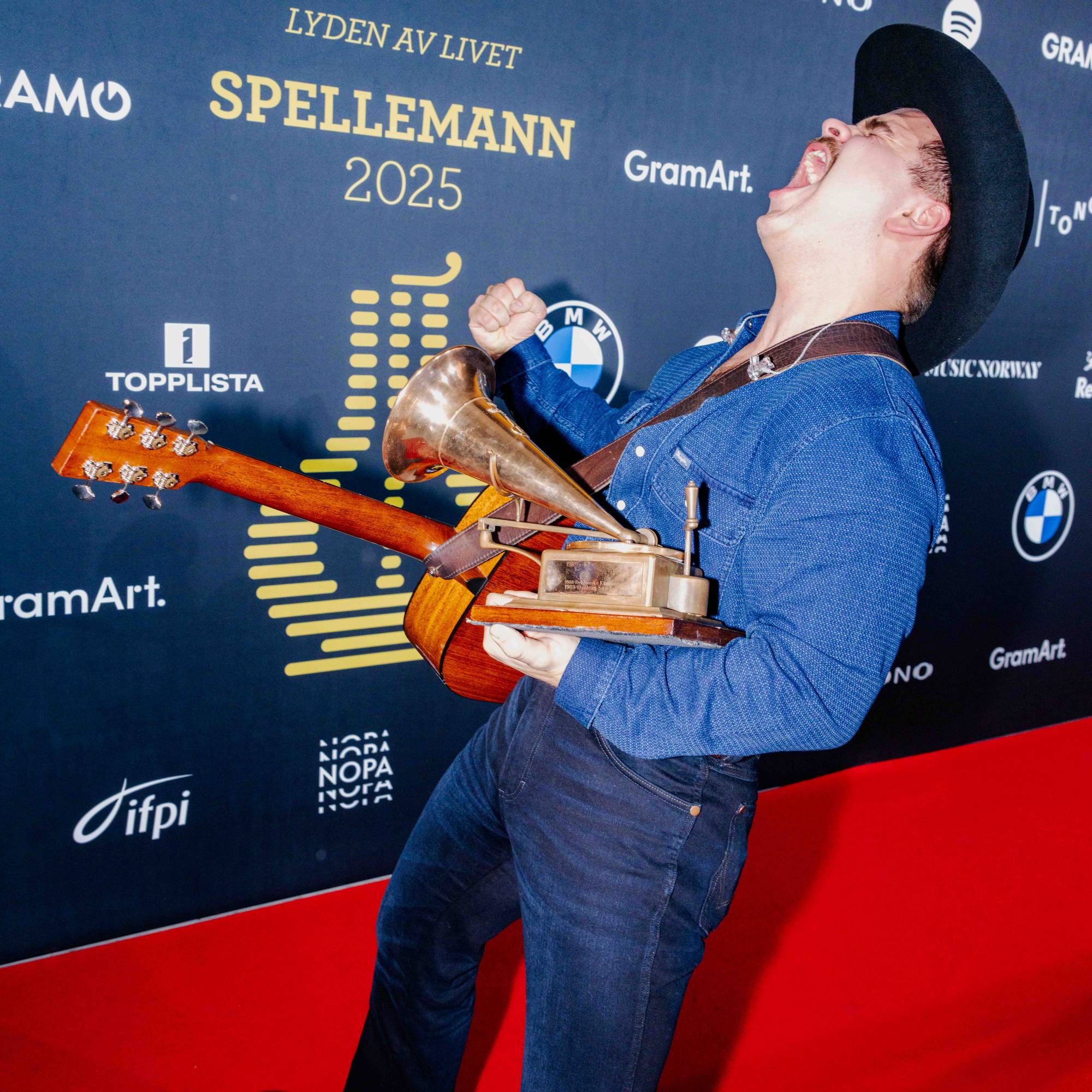 En mann i cowboyhatt feirer med gitar og pris på rød løper. Mannen har en blå skjorte og mørke jeans. Bakgrunnen er en mørkeblå vegg med logoer. (Bildebeskrivelsen er laget av en KI-tjeneste)