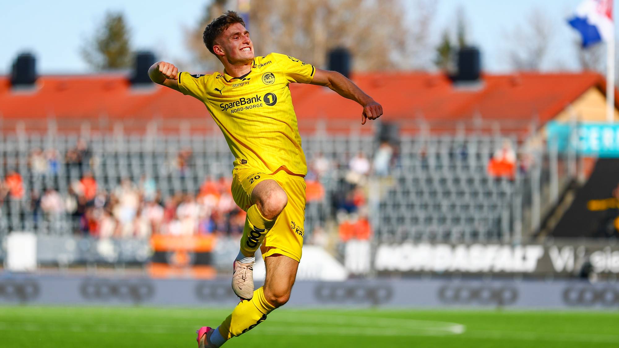En fotballspiller i gul drakt jubler på en gressbane. Han er i luften, med armen hevet. I bakgrunnen er det en tribune med tilskuere og et flagg. Solen skinner. (Bildebeskrivelsen er laget av en KI-tjeneste)