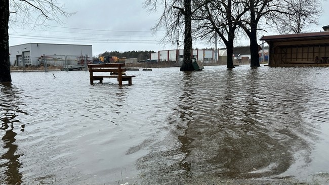 Bildet viser en oversvømt område med en benk som står halvveis nedsenket i vannet. Det er flere trær på motsatt side av benken, og i bakgrunnen kan man skimte industrielle bygninger og gjerder. Vannflaten er rolig og reflekterer omgivelsene. Området synes å være rammet av betydelig nedbør. (Bildebeskrivelsen er laget av en KI-tjeneste)