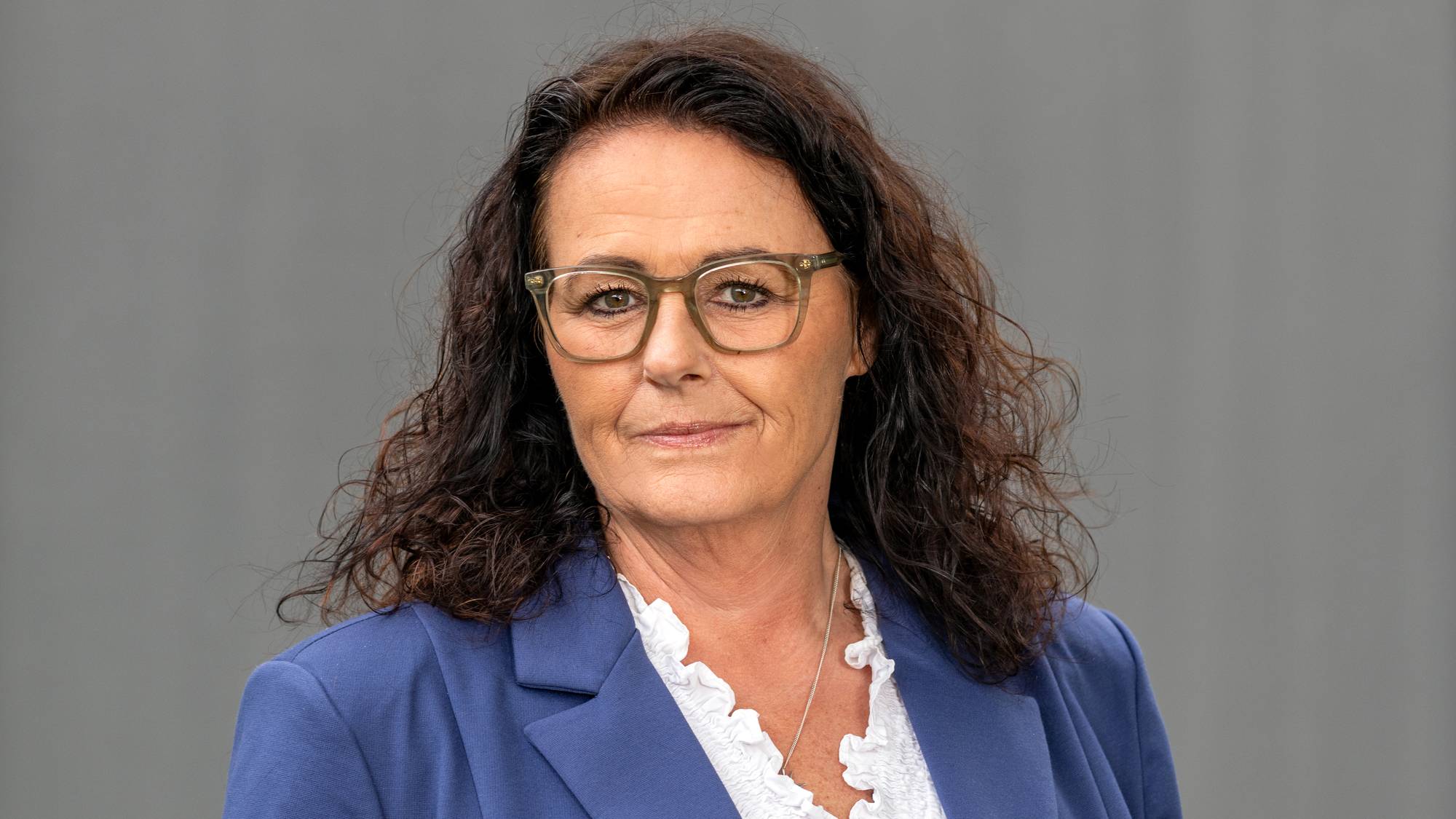 Kvinne med mørkt hår og briller, kledd i blå blazer og hvit bluse, foran en grå bakgrunn. (Bildebeskrivelsen er laget av en KI-tjeneste)